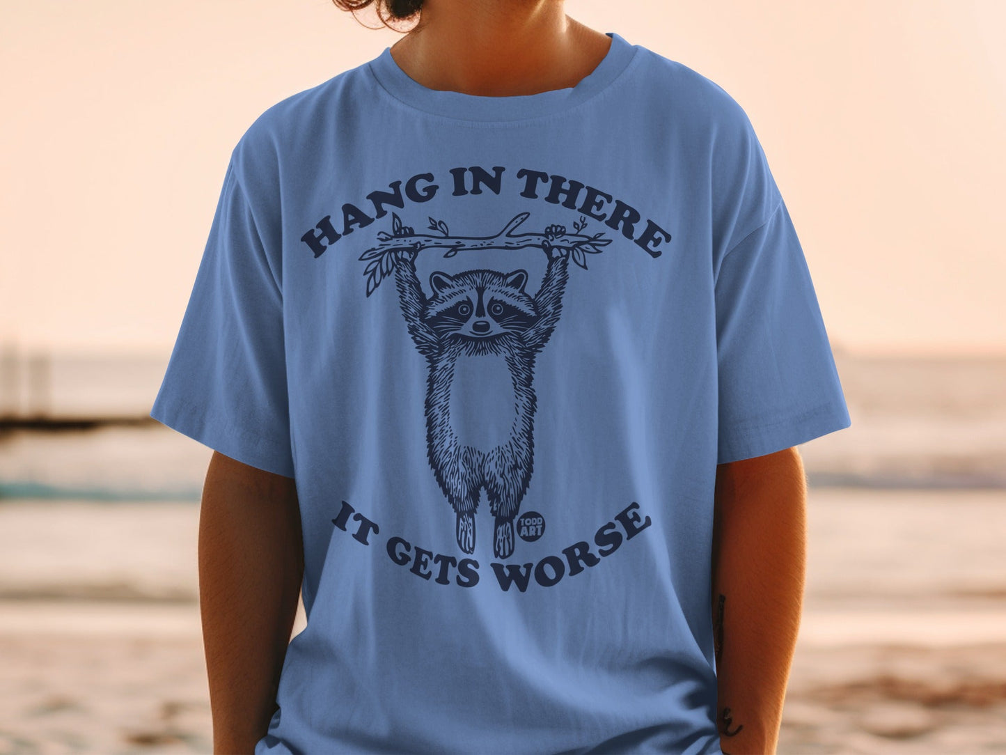 Blue t-shirt with a raccoon hanging from a branch, text says "HANG IN THERE IT GETS WORSE".