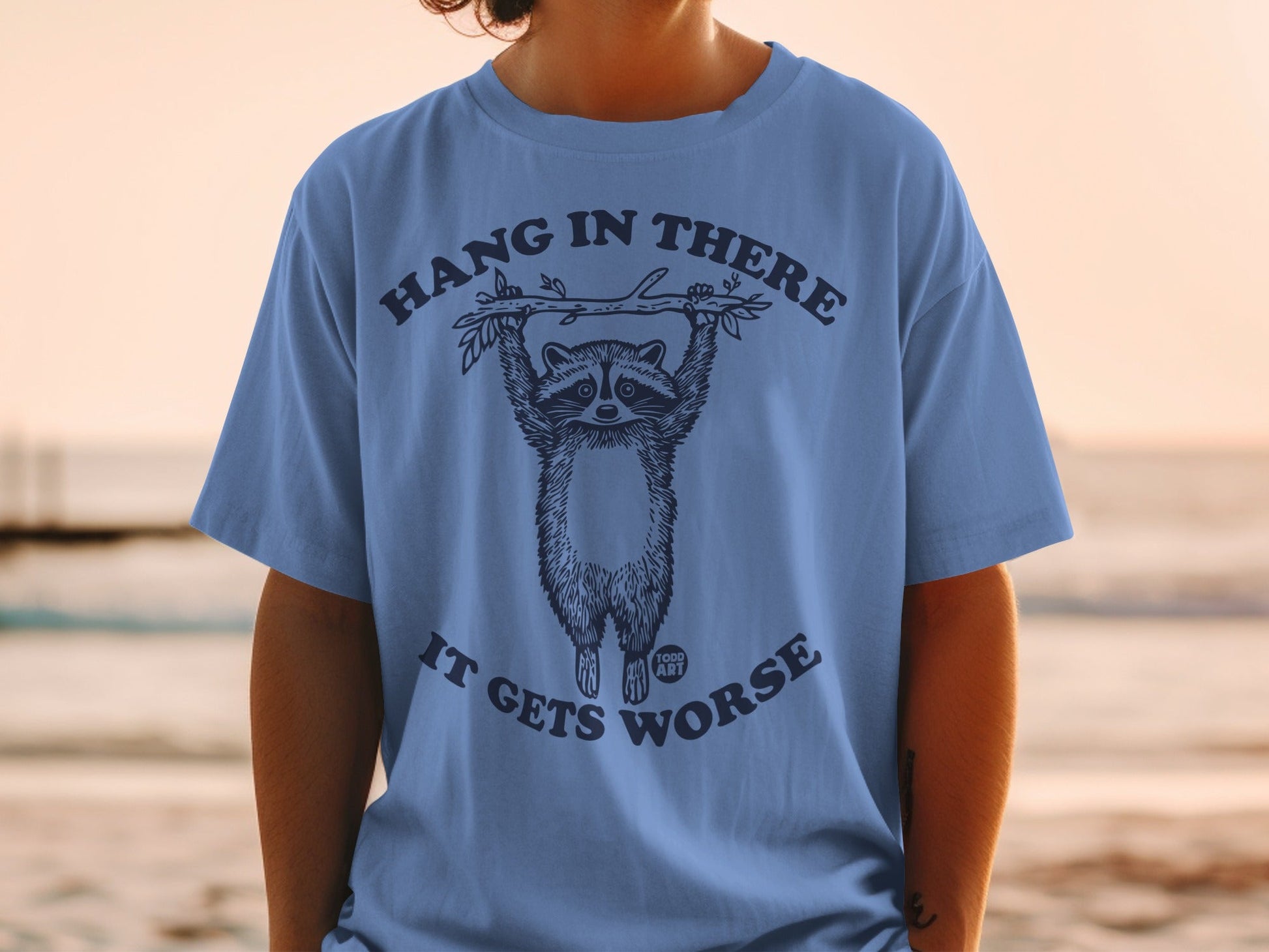 Blue t-shirt with a raccoon hanging from a branch, text says "HANG IN THERE IT GETS WORSE".