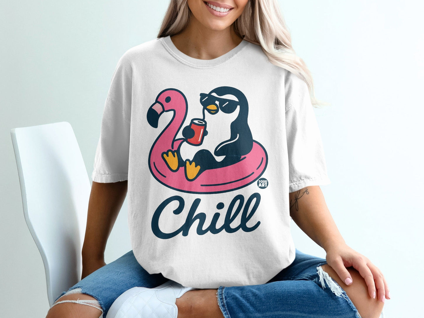 White t-shirt with a cartoon penguin on a flamingo float, holding a drink.