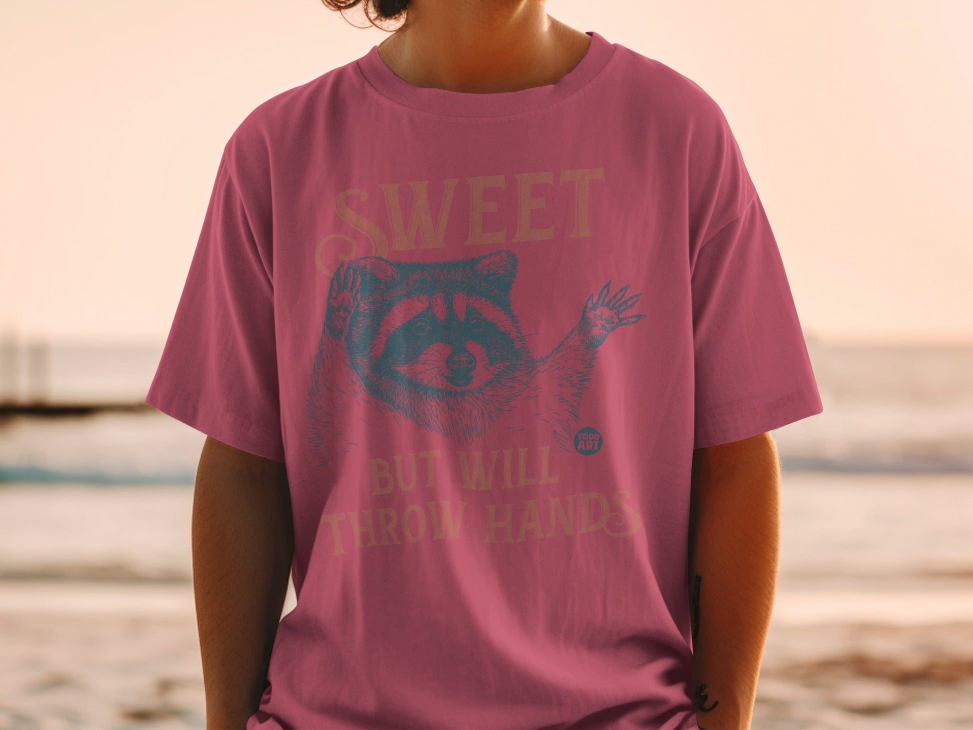 Pink t-shirt with raccoon illustration and text "SWEET BUT WILL THROW HANDS".