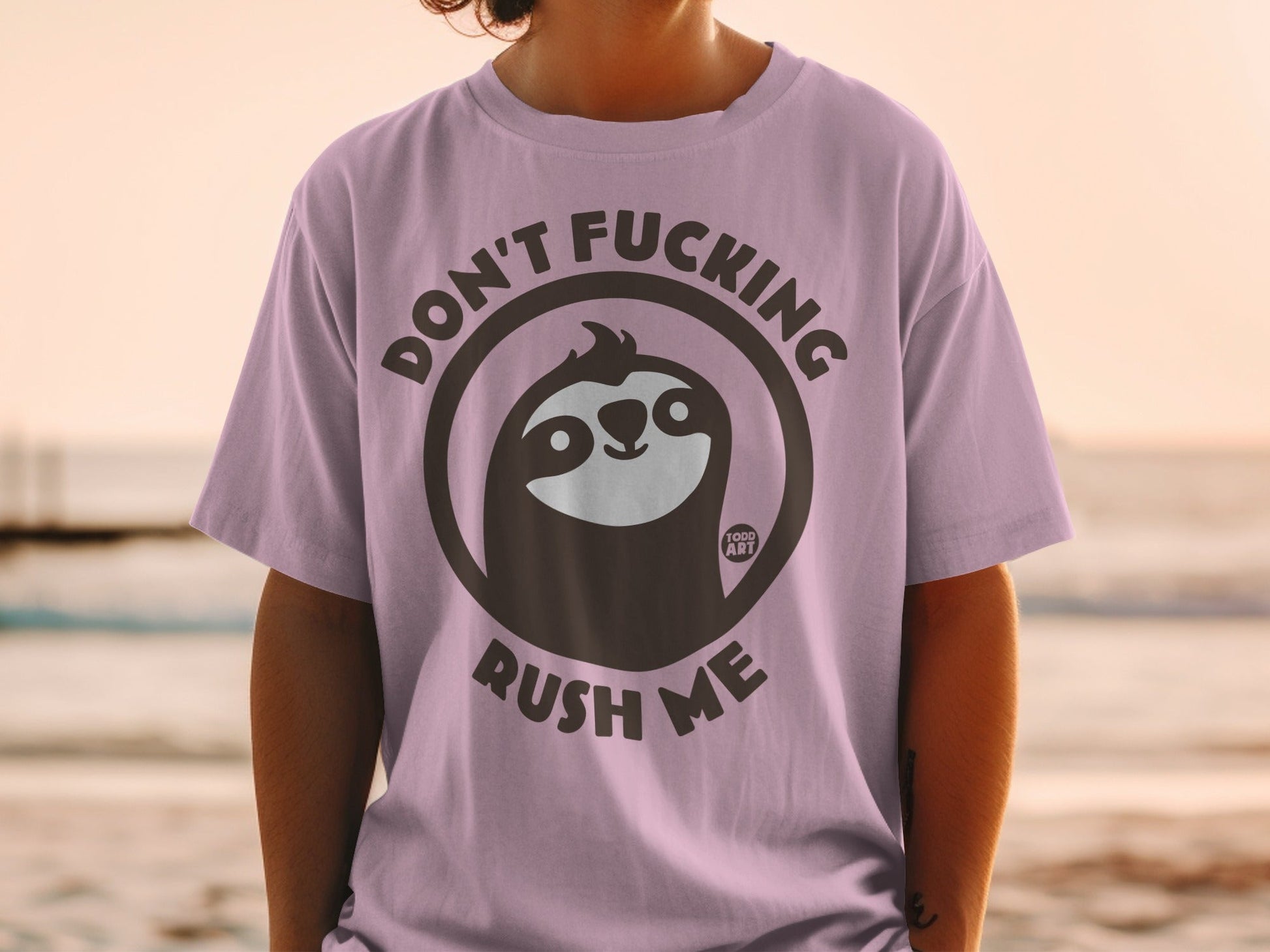 Sloth graphic with "Don't Fucking Rush Me" text on a light purple t-shirt.