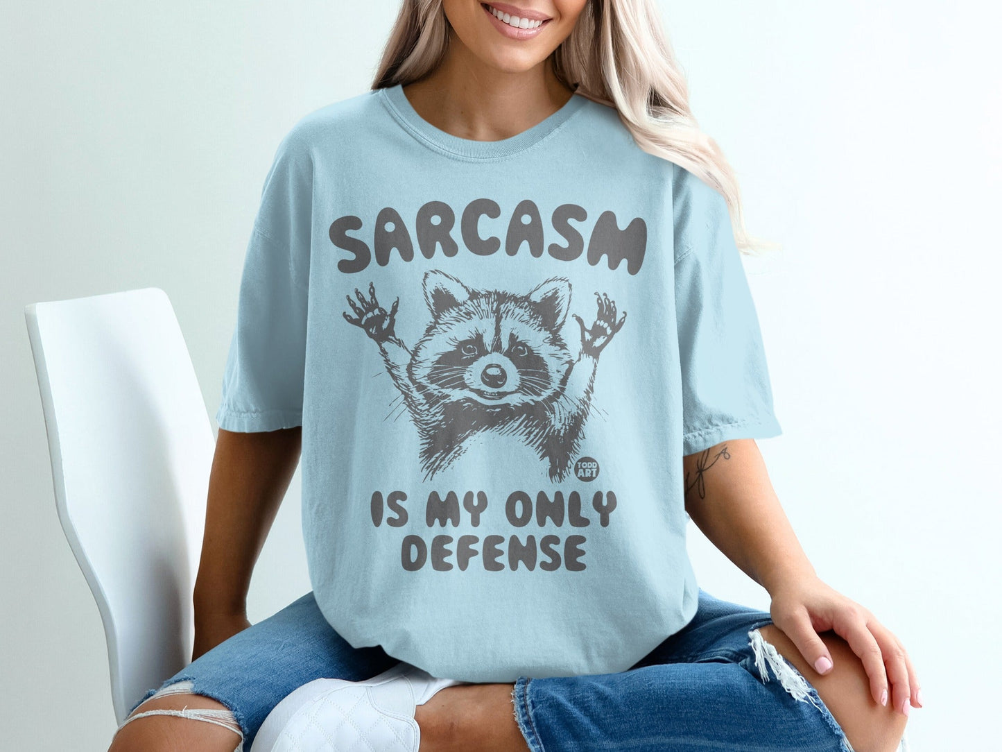 Light blue t-shirt with raccoon graphic and "SARCASM IS MY ONLY DEFENSE" text.