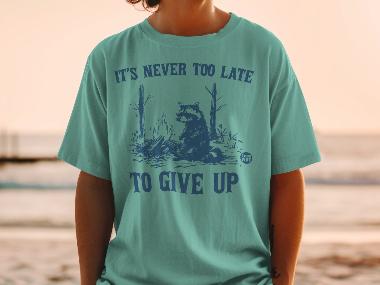 Teal t-shirt with raccoon by campfire graphic and "It's never too late to give up" text.