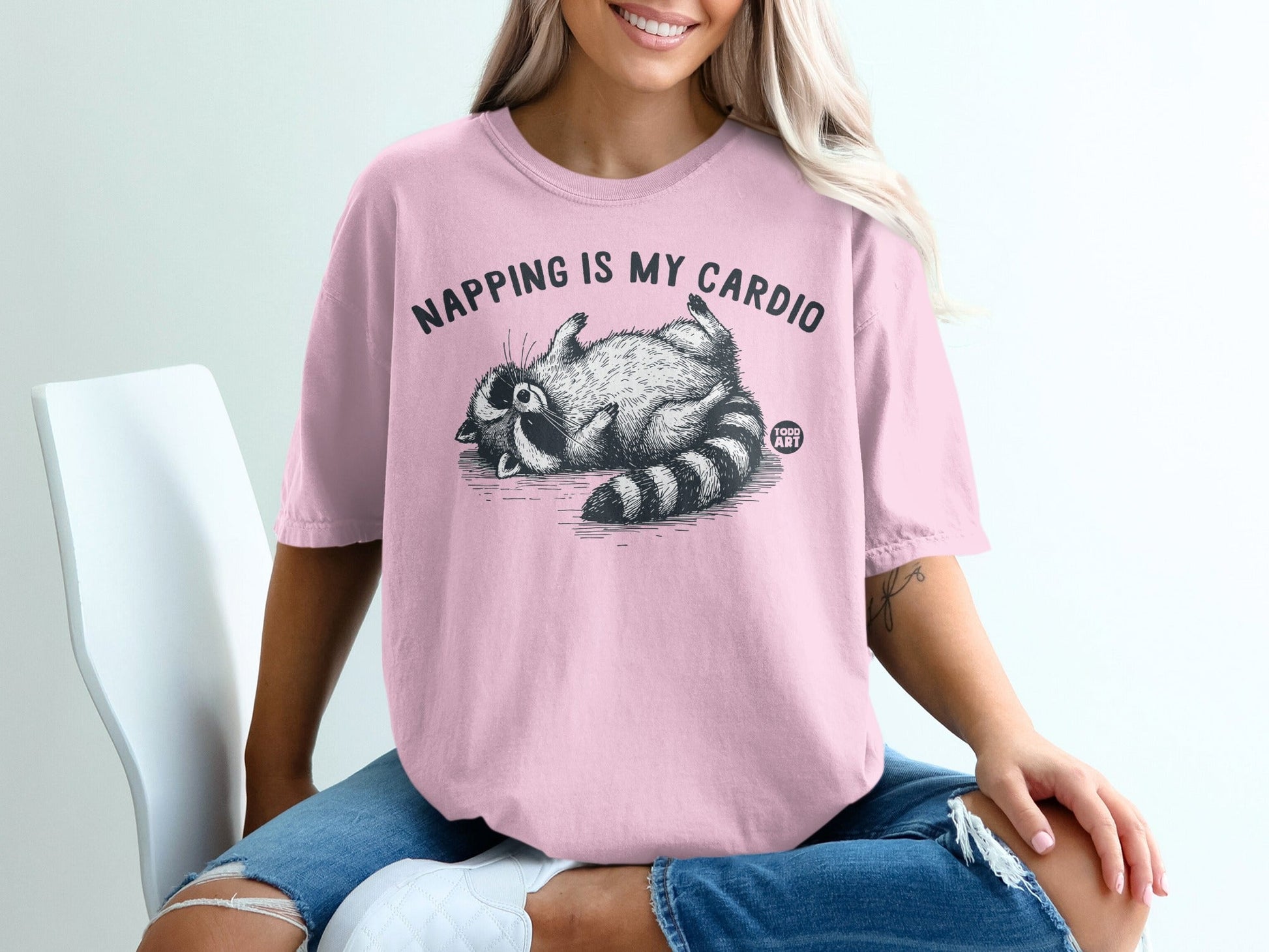 Pink t-shirt with a graphic of a raccoon lounging and the text "Napping is my cardio".