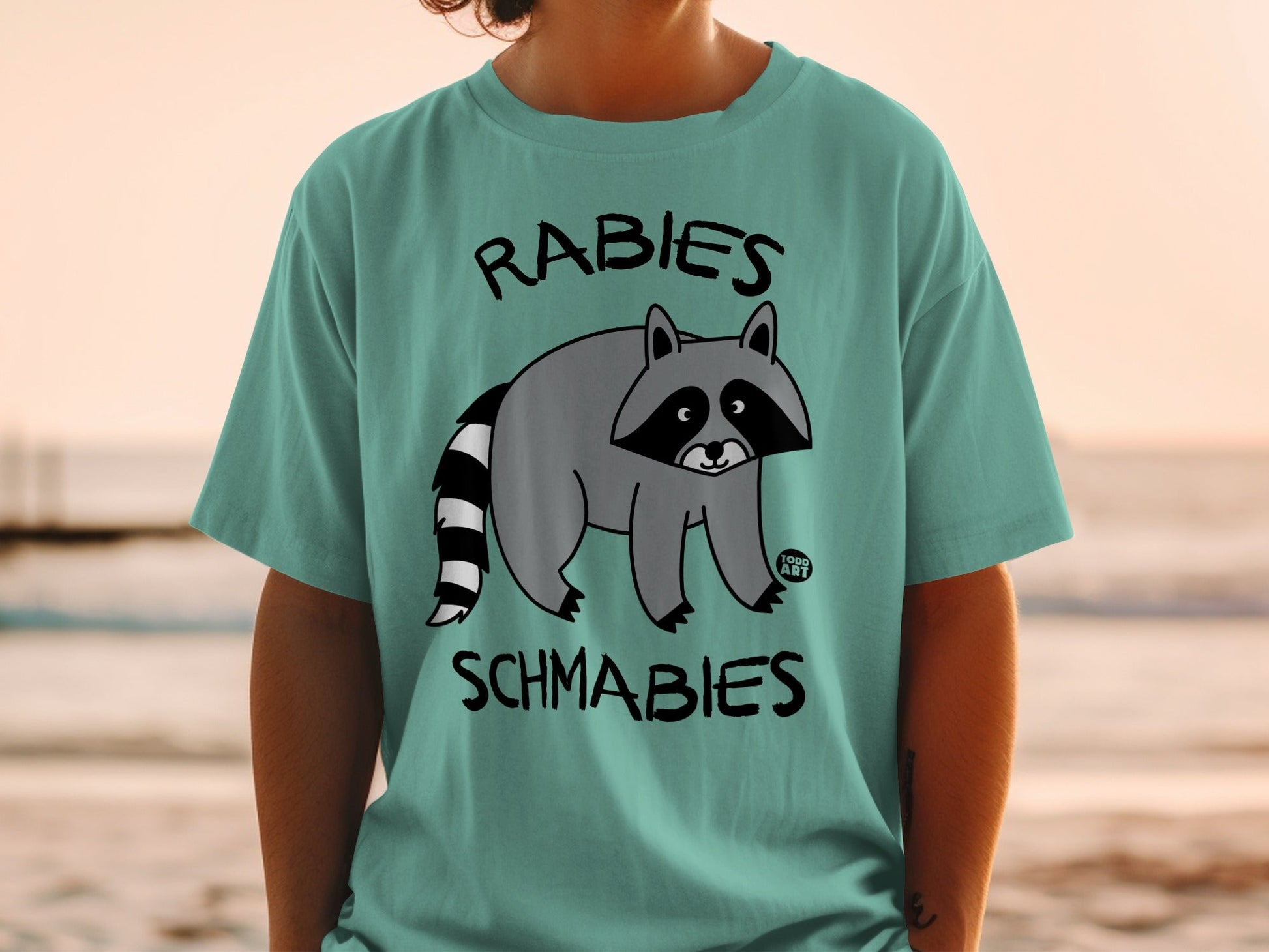 Teal t-shirt with a cartoon raccoon and text "RABIES SCHMABIES".