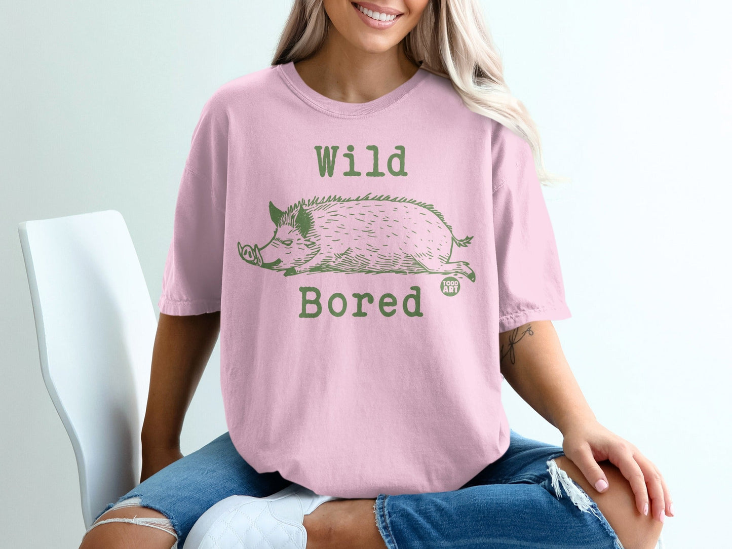 Pink t-shirt with a green illustration of a sleeping wild boar and "Wild Bored" text.
