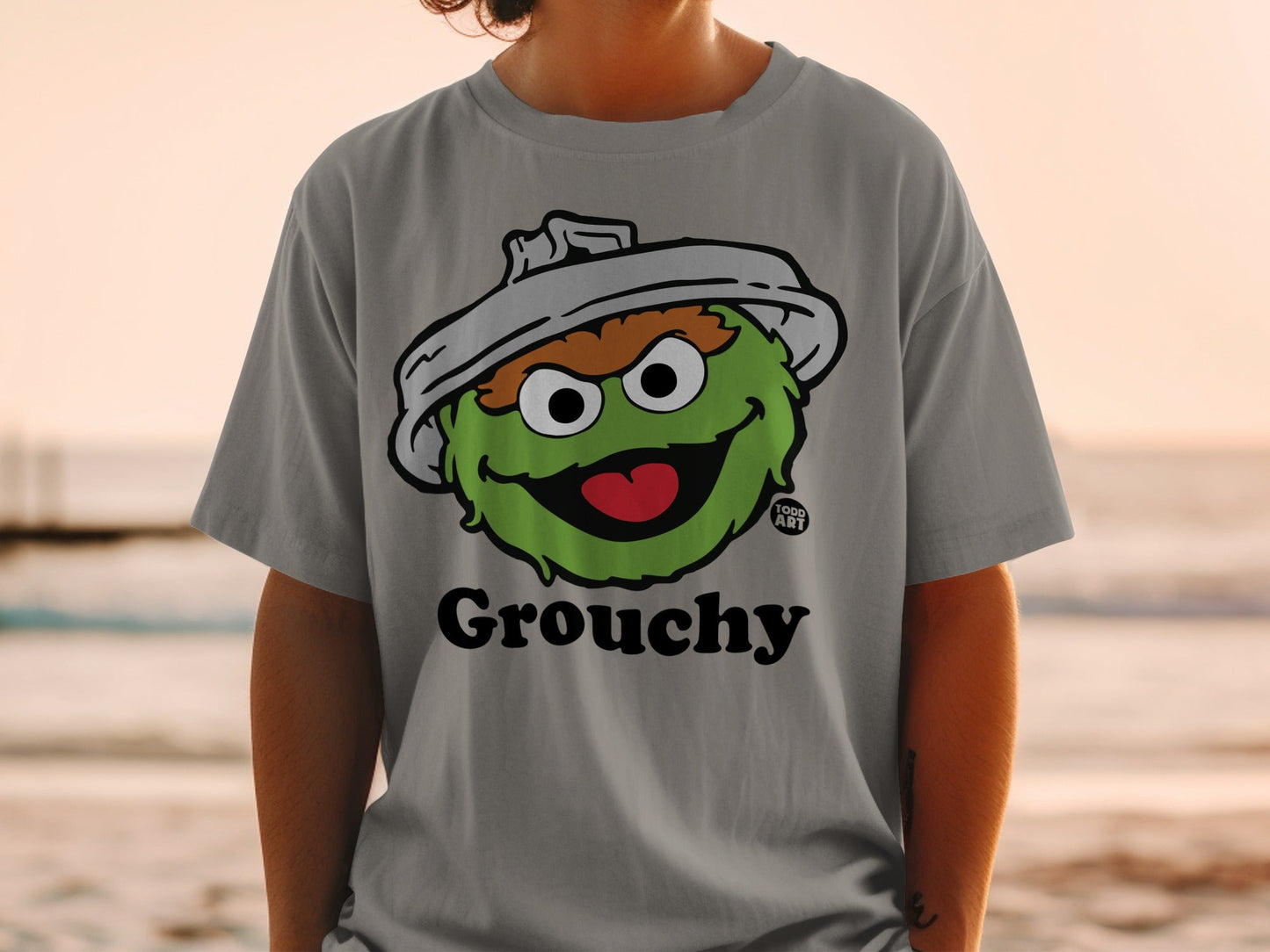 Grey t-shirt featuring a cartoon green monster with a trash can lid hat and the word "Grouchy."
