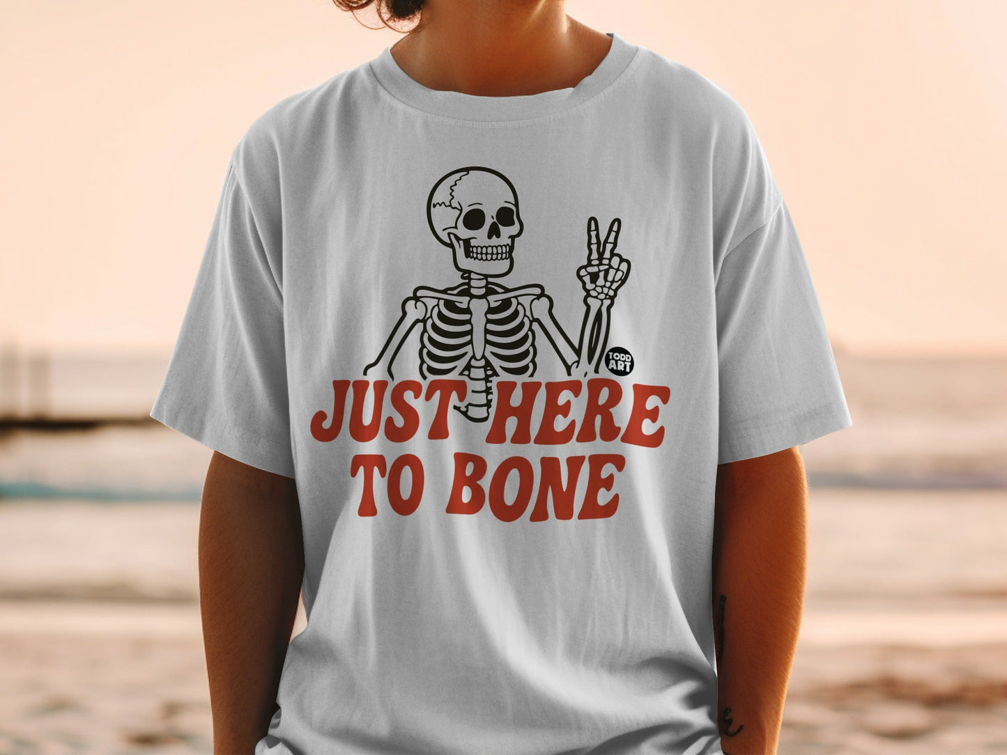 White t-shirt with skeleton making peace sign and text "JUST HERE TO BONE".