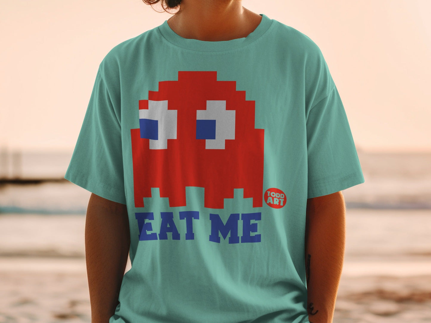Red Pac-Man ghost graphic with "EAT ME" text on a teal t-shirt.