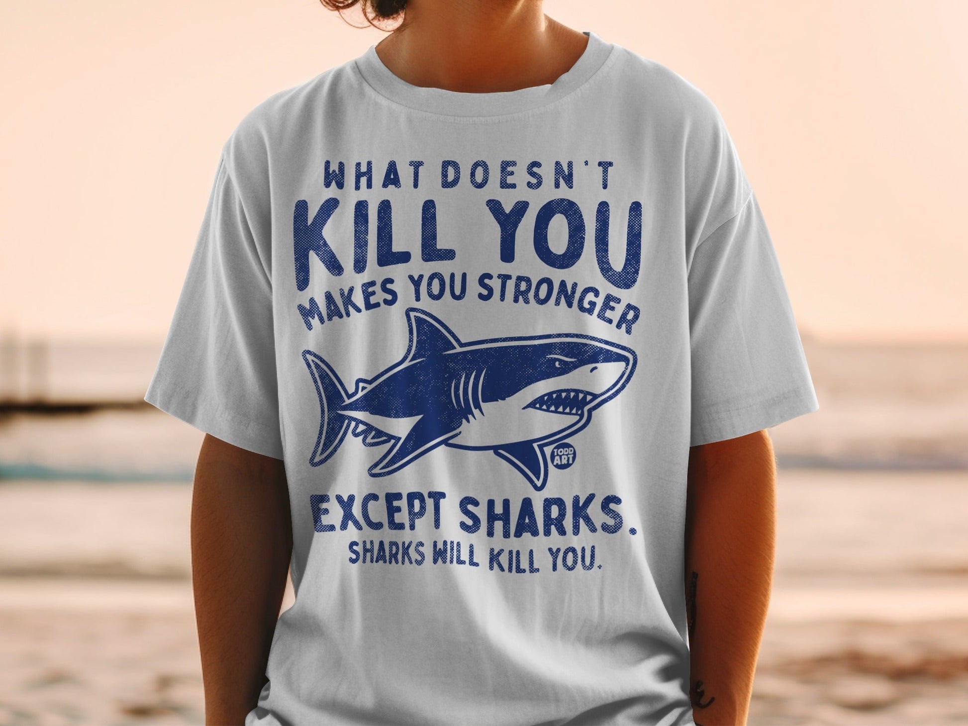 T-shirt with shark graphic and text: "What doesn't kill you makes you stronger, except sharks."