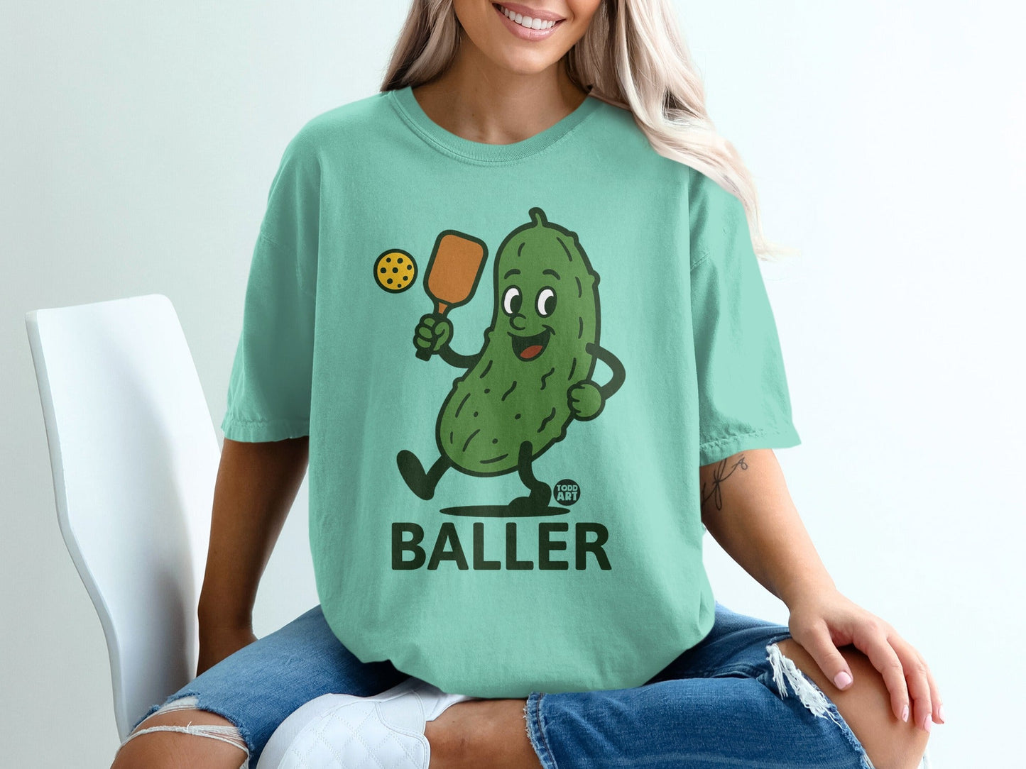 Cartoon pickle wearing a shirt, holding a paddle and pickleball.