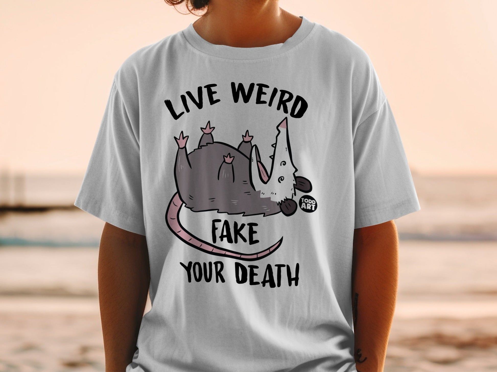 White t-shirt with possum graphic and text "LIVE WEIRD FAKE YOUR DEATH".