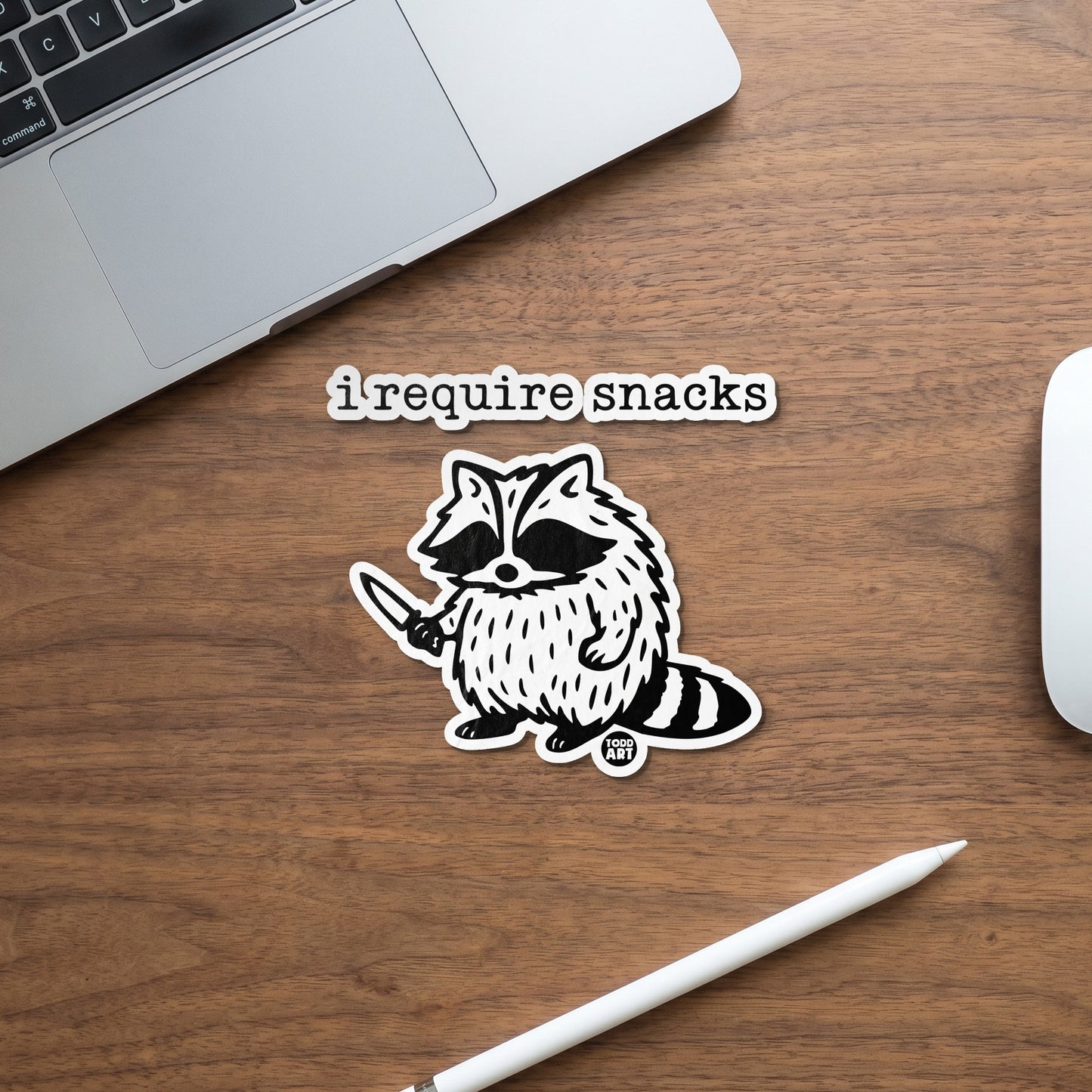 Black and white raccoon sticker holding a knife with text "i require snacks".