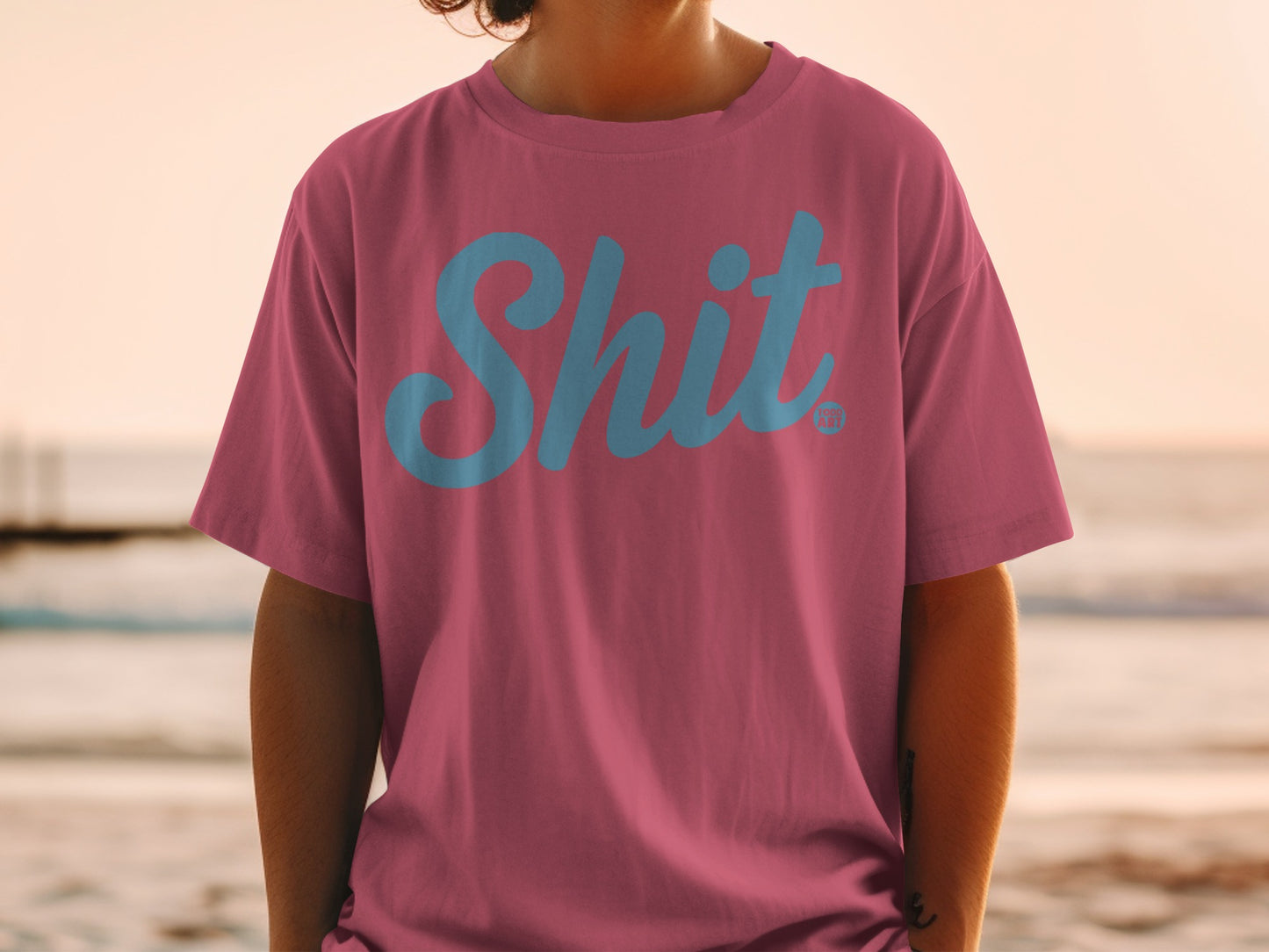 Pink t-shirt with blue "Shit" logo, worn by a person at the beach.
