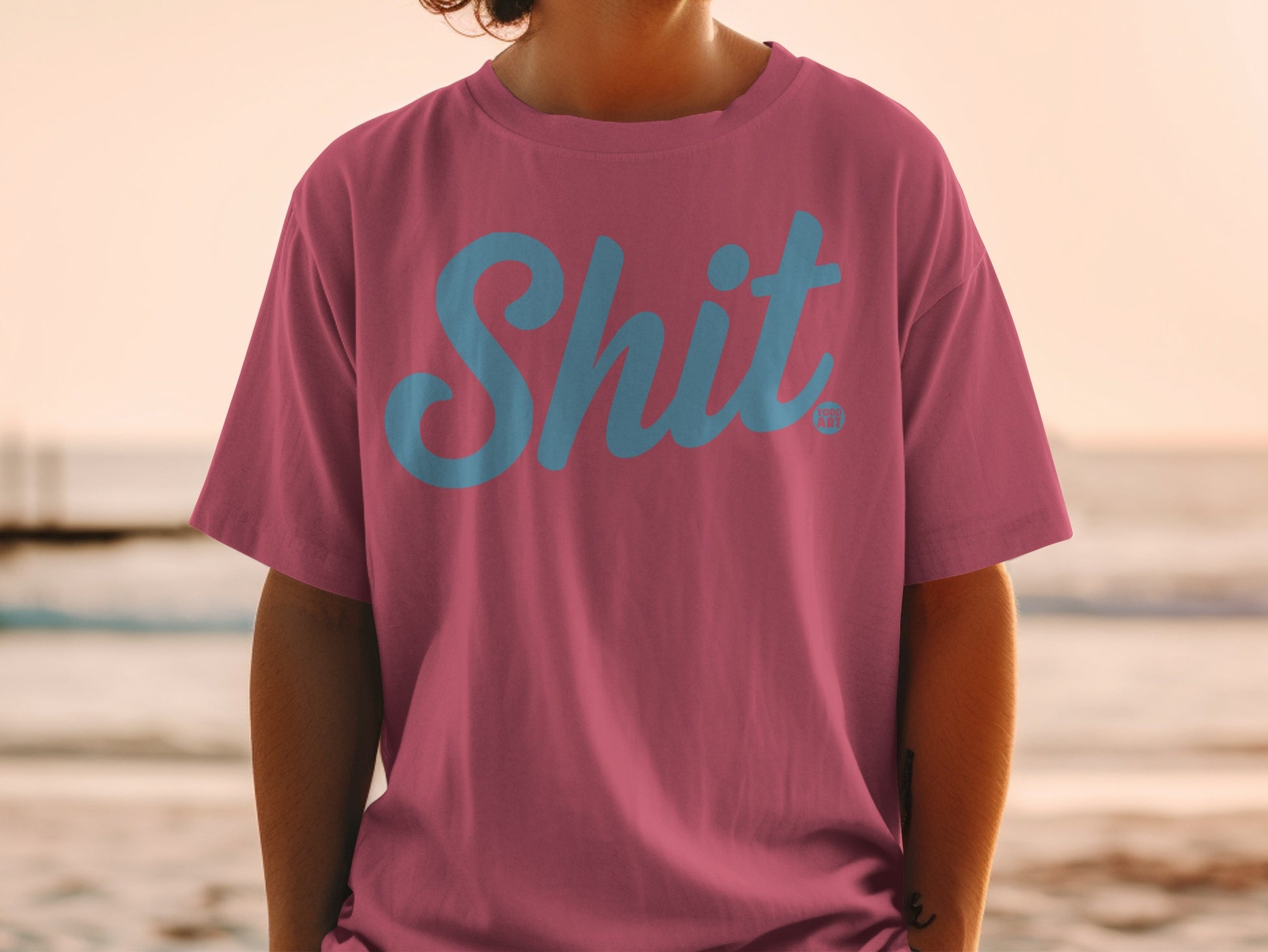Pink t-shirt with blue "Shit" logo, worn by a person at the beach.