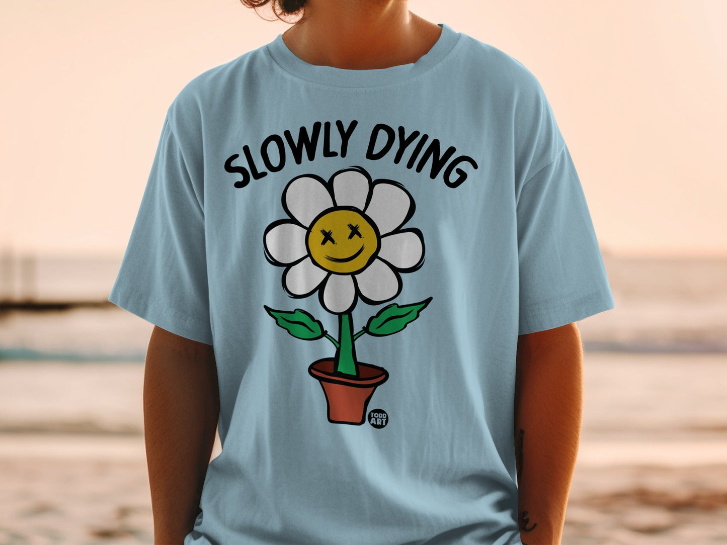 Blue t-shirt with a smiling, dead-eyed flower in a pot, reading "SLOWLY DYING".