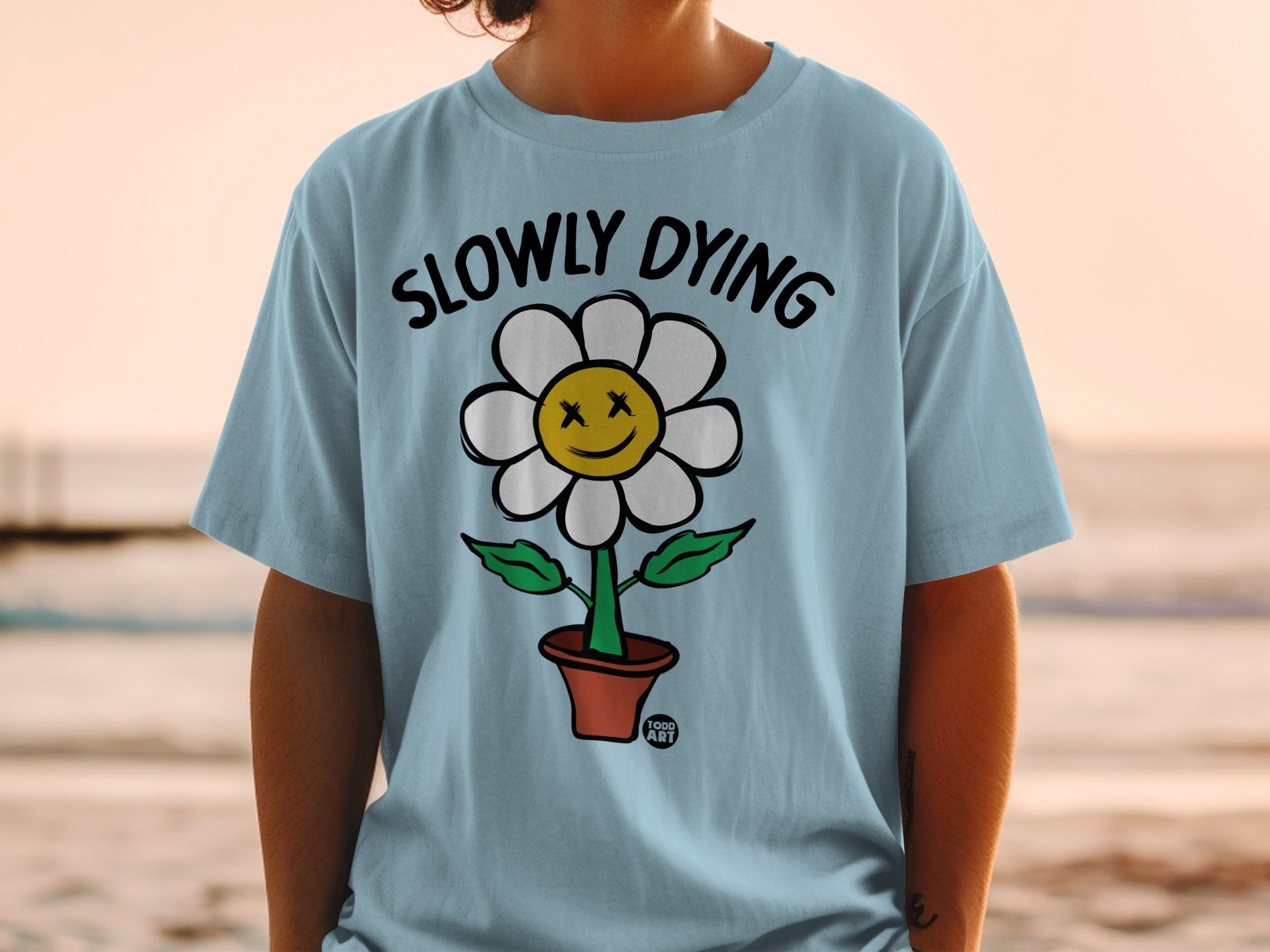 Blue t-shirt with a smiling, dead-eyed flower in a pot, reading "SLOWLY DYING".