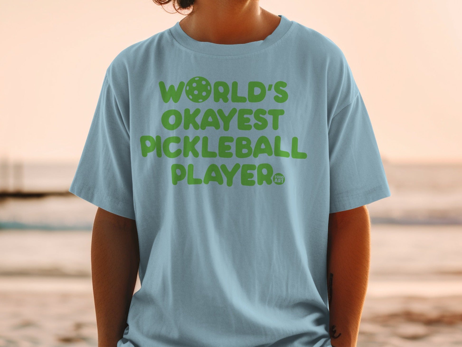 Light blue t-shirt with "World's Okayest Pickleball Player" text in green.