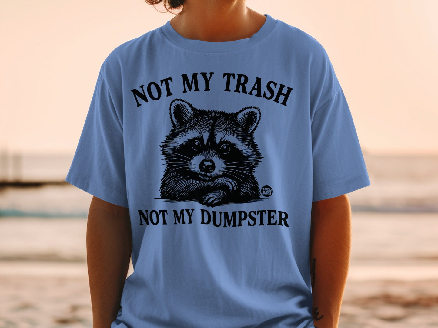 Blue t-shirt with raccoon illustration and "NOT MY TRASH, NOT MY DUMPSTER" text.