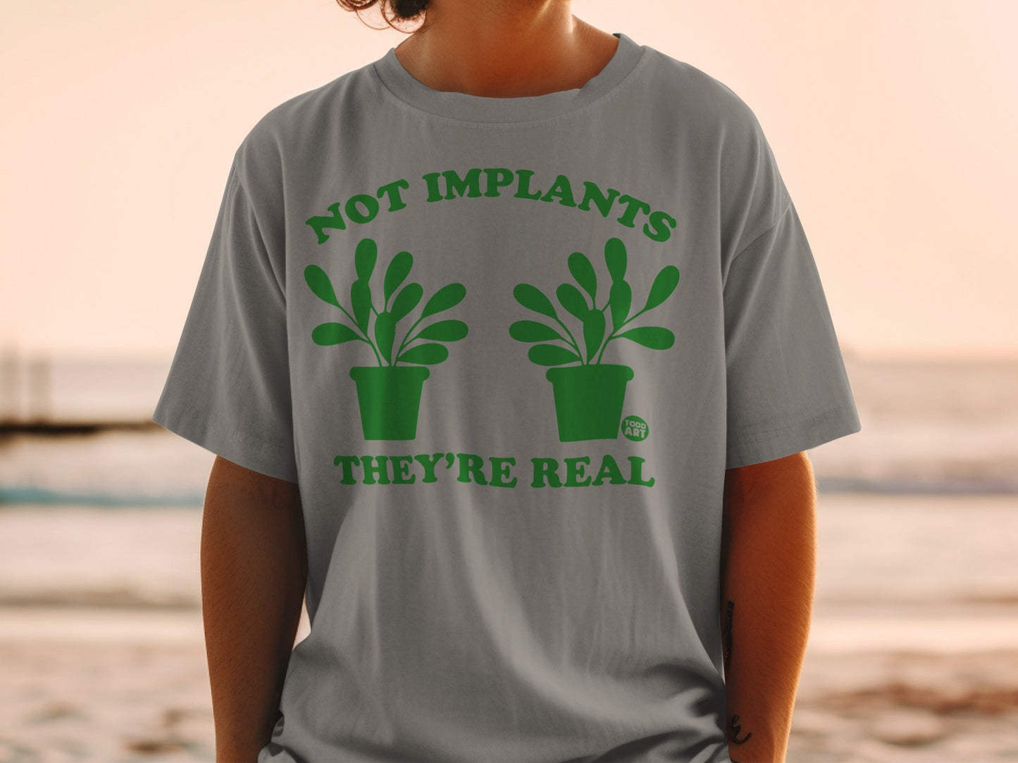 Grey t-shirt with green text and potted plants: "NOT IMPLANTS THEY'RE REAL"