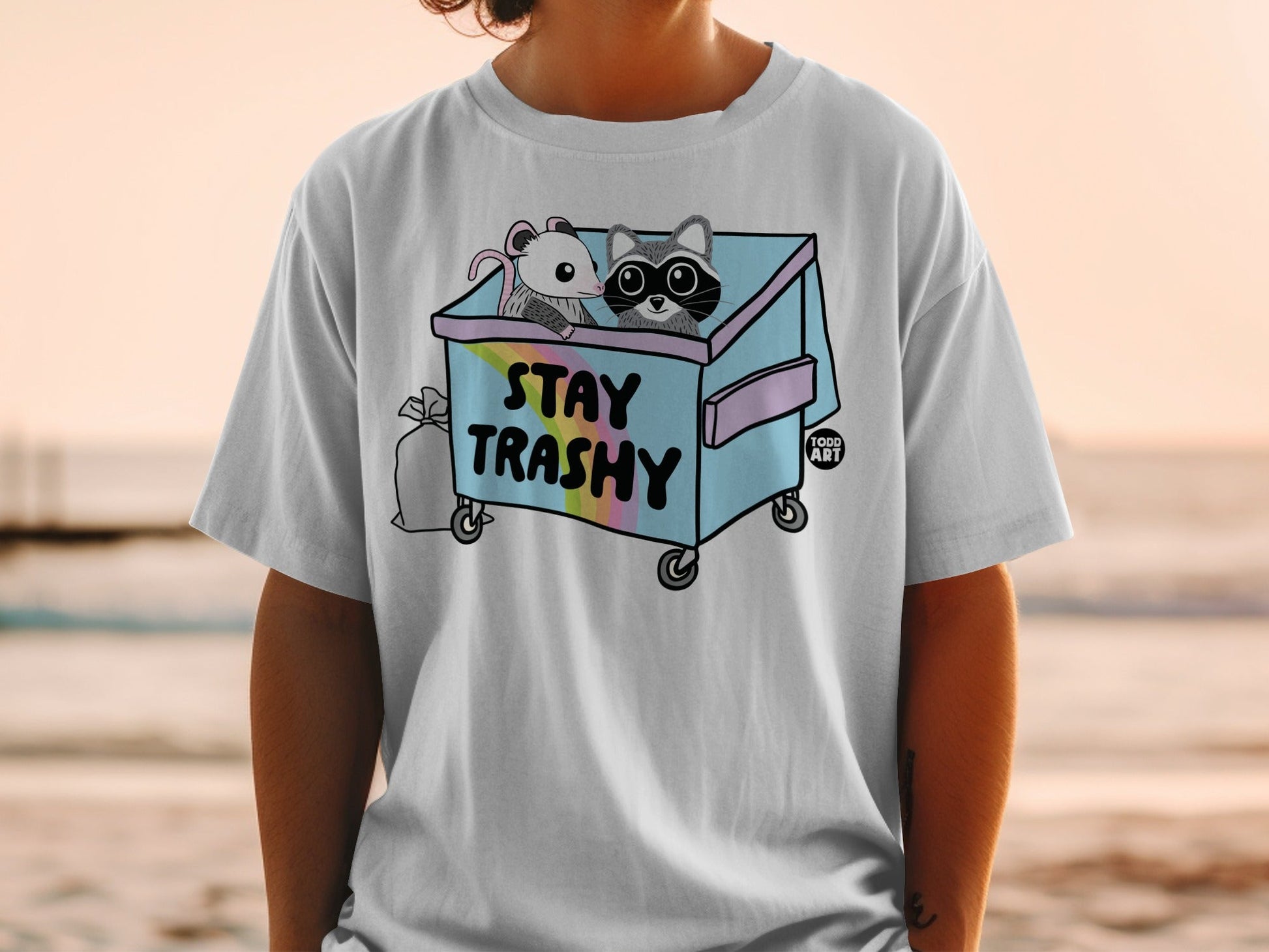 T-shirt graphic of a raccoon and possum in a pastel trash can with "STAY TRASHY" text.