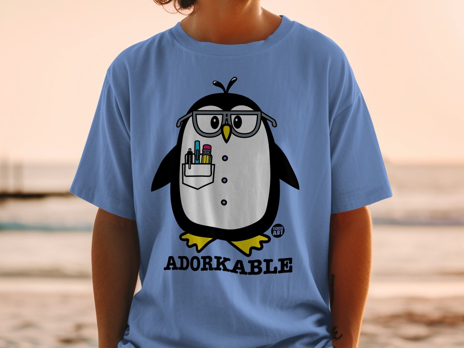 Light blue t-shirt with a cartoon penguin wearing glasses holding pens.