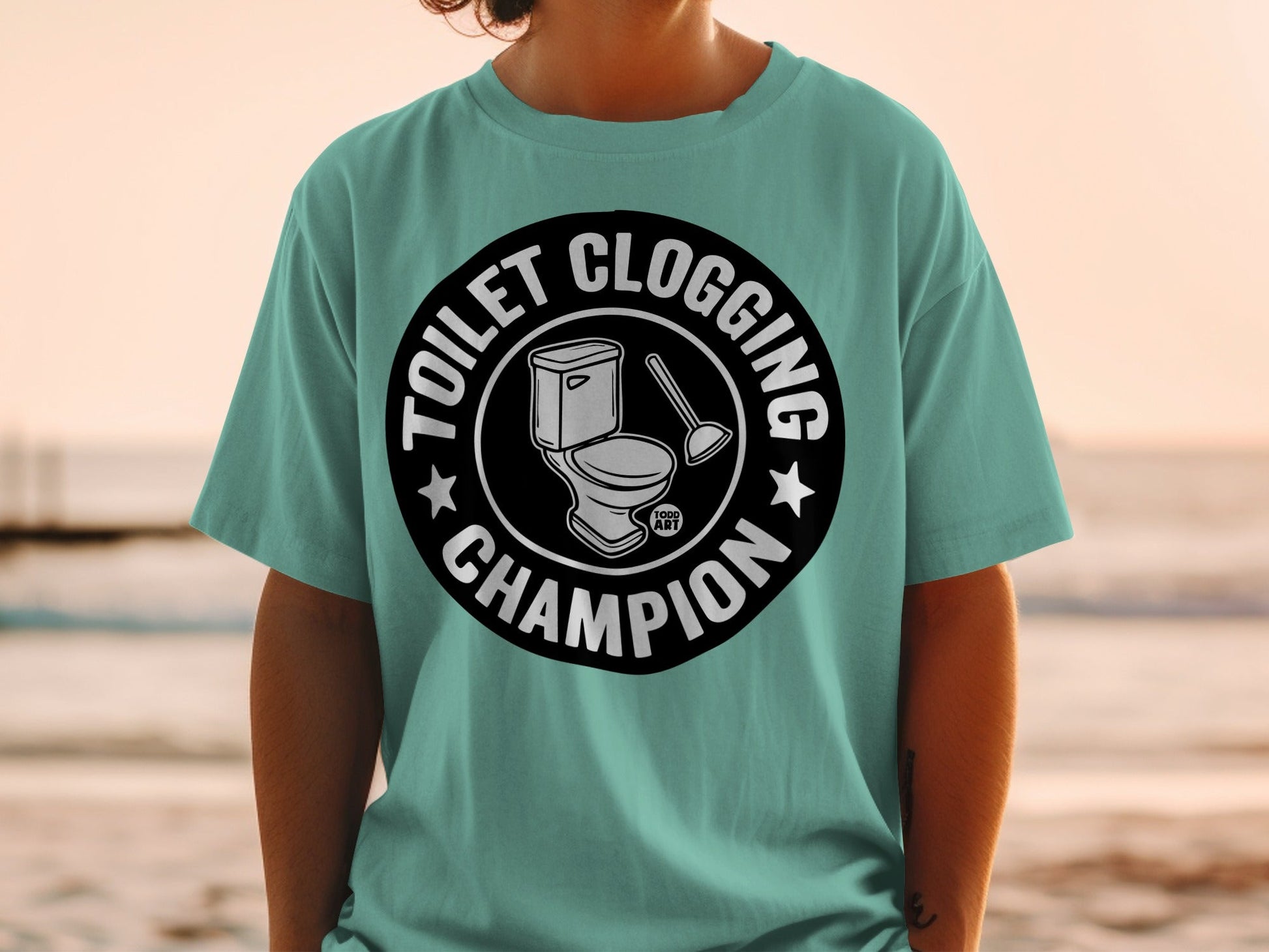 Teal t-shirt with circular graphic of a toilet and plunger, reading "Toilet Clogging Champion".