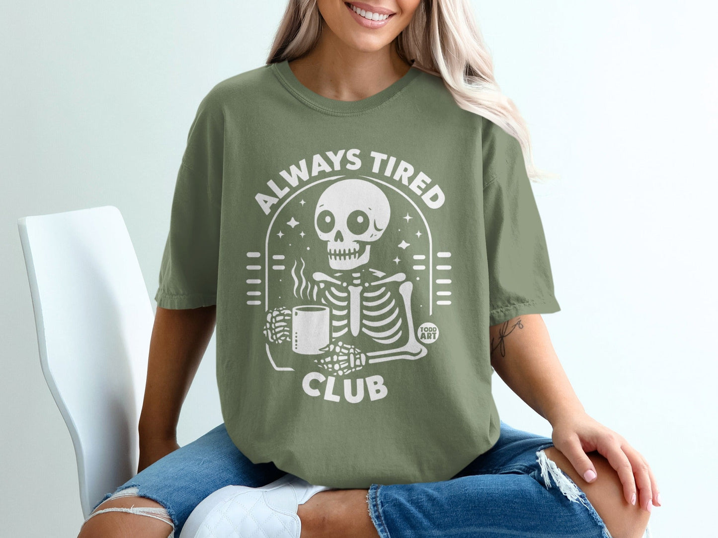 Skeleton holding coffee mug with "Always Tired Club" text on olive green t-shirt.