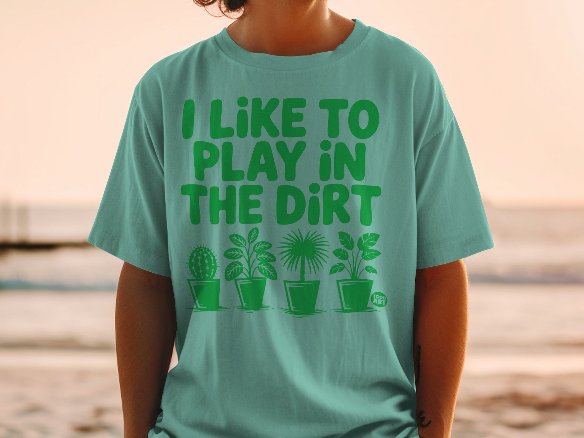 Teal t-shirt with green text "I LIKE TO PLAY IN THE DIRT" and potted plants.