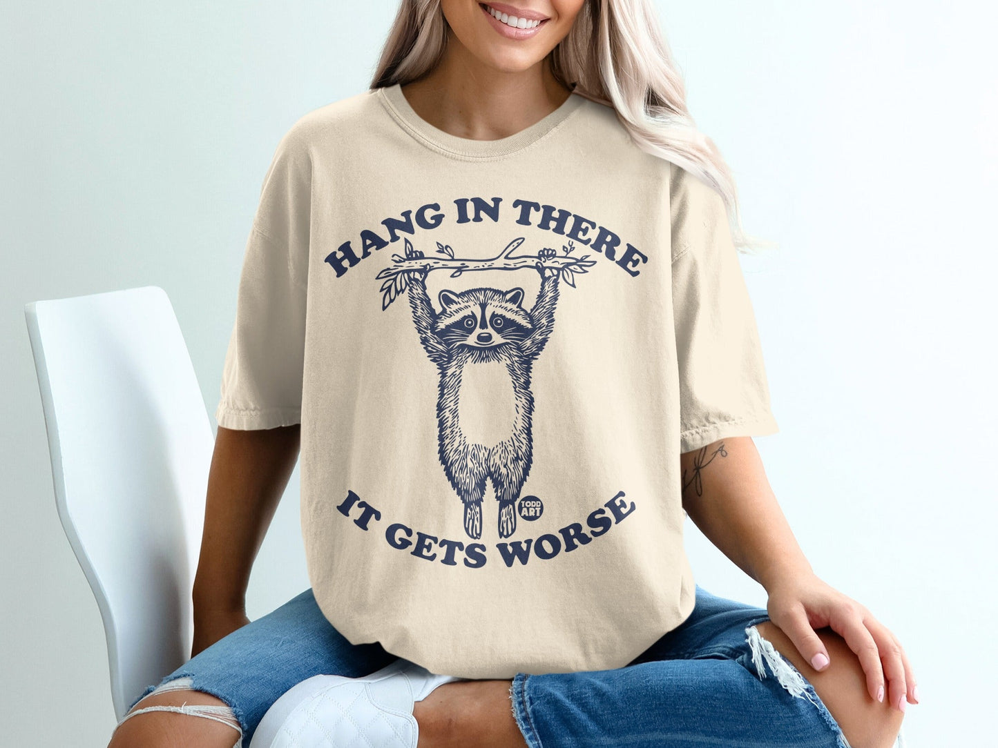 T-shirt with raccoon hanging from branch, text reads "HANG IN THERE IT GETS WORSE."