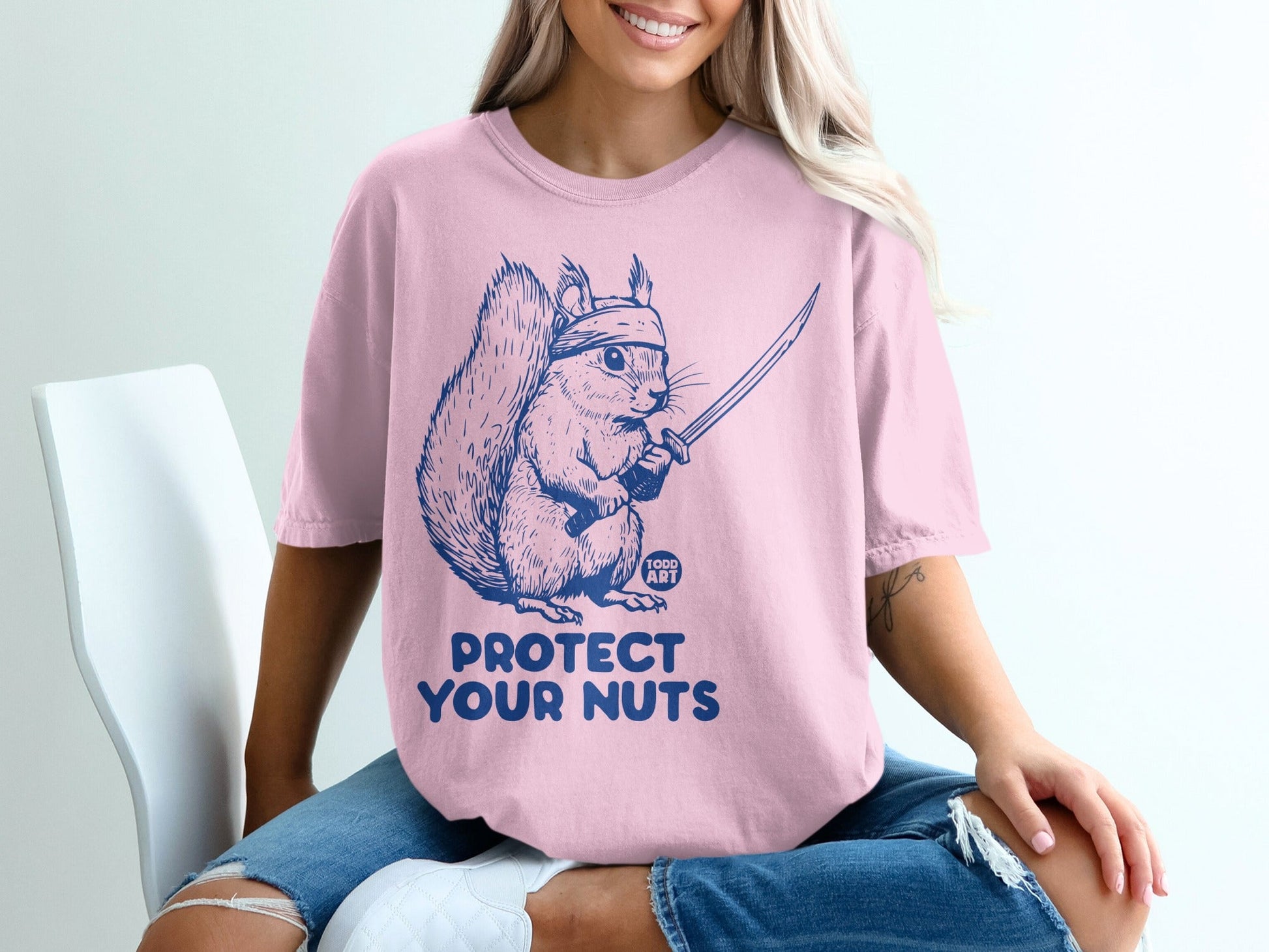 Pink t-shirt with blue graphic of a squirrel holding a sword and the text "PROTECT YOUR NUTS".