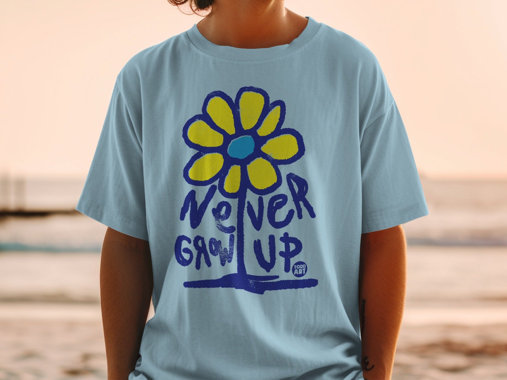 Light blue t-shirt with a yellow flower and "Never Grow Up" text.