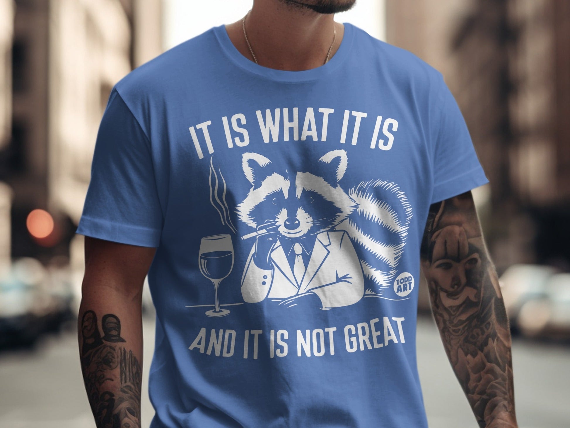 Blue t-shirt with raccoon in suit, smoking and holding wine glass. Text reads "It is what it is and it is not great."