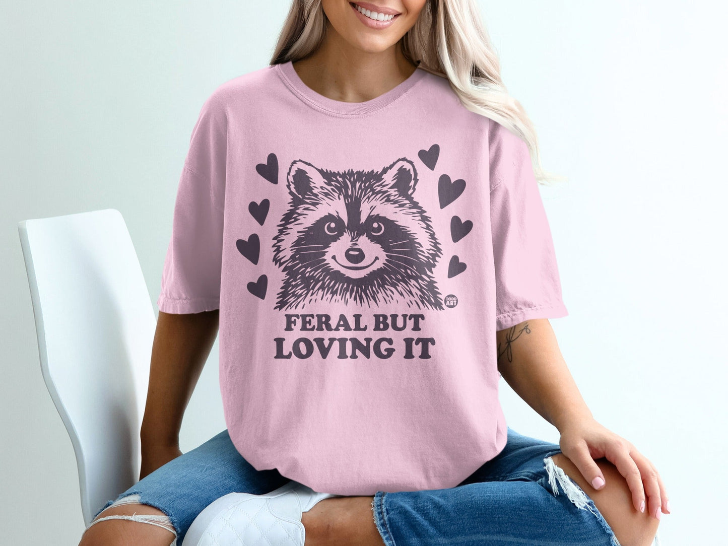 Pink t-shirt with a raccoon and hearts that reads "FERAL BUT LOVING IT."