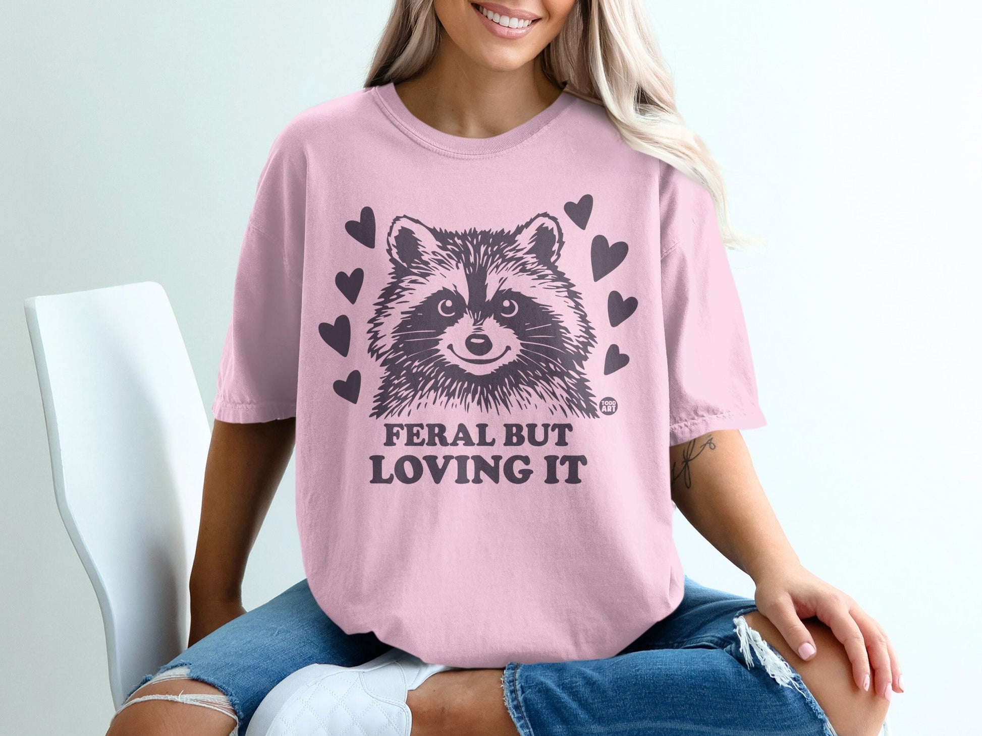 Pink t-shirt with a raccoon and hearts that reads "FERAL BUT LOVING IT."