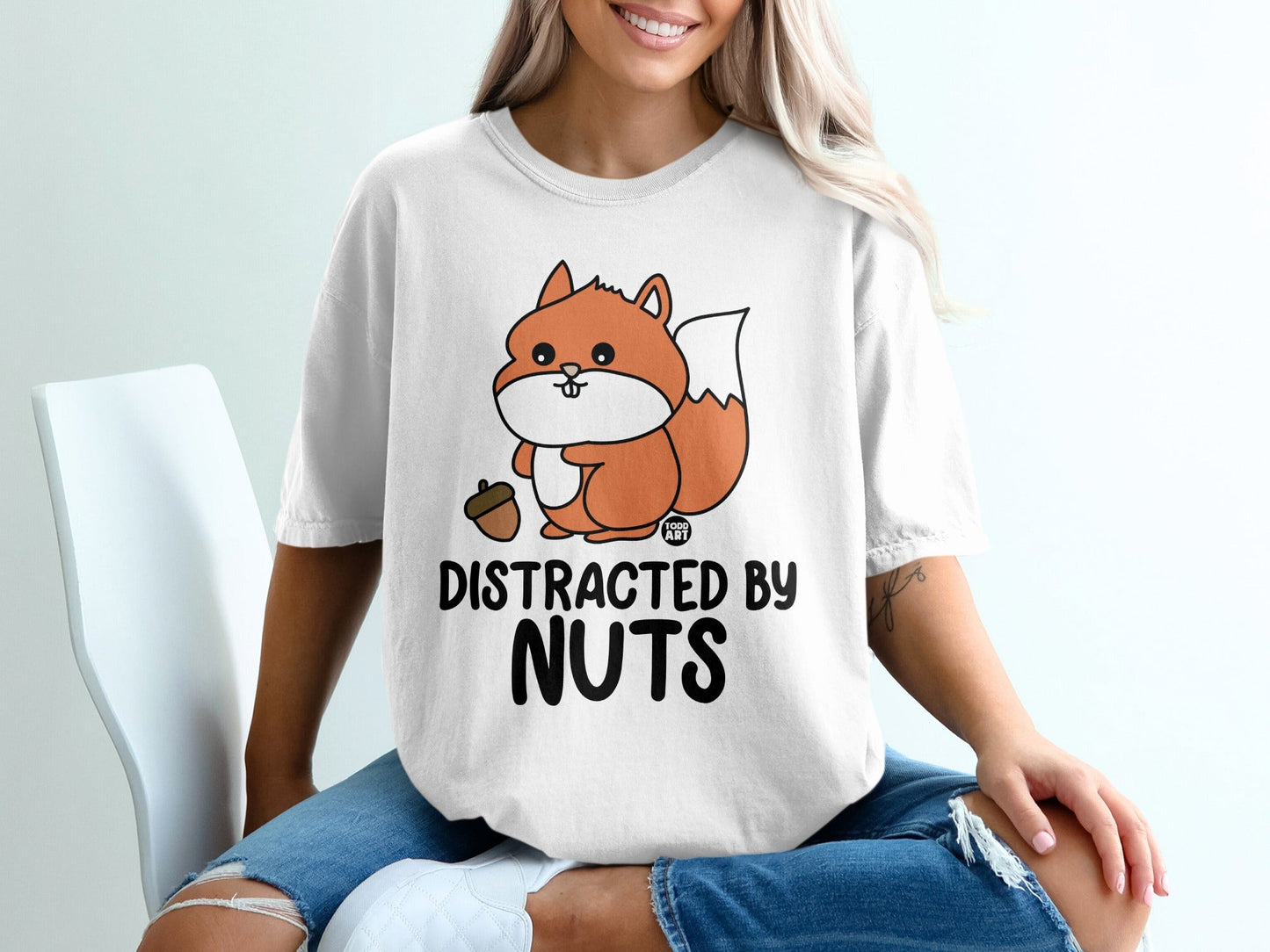 White t-shirt with a cartoon squirrel holding an acorn, text "DISTRACTED BY NUTS".