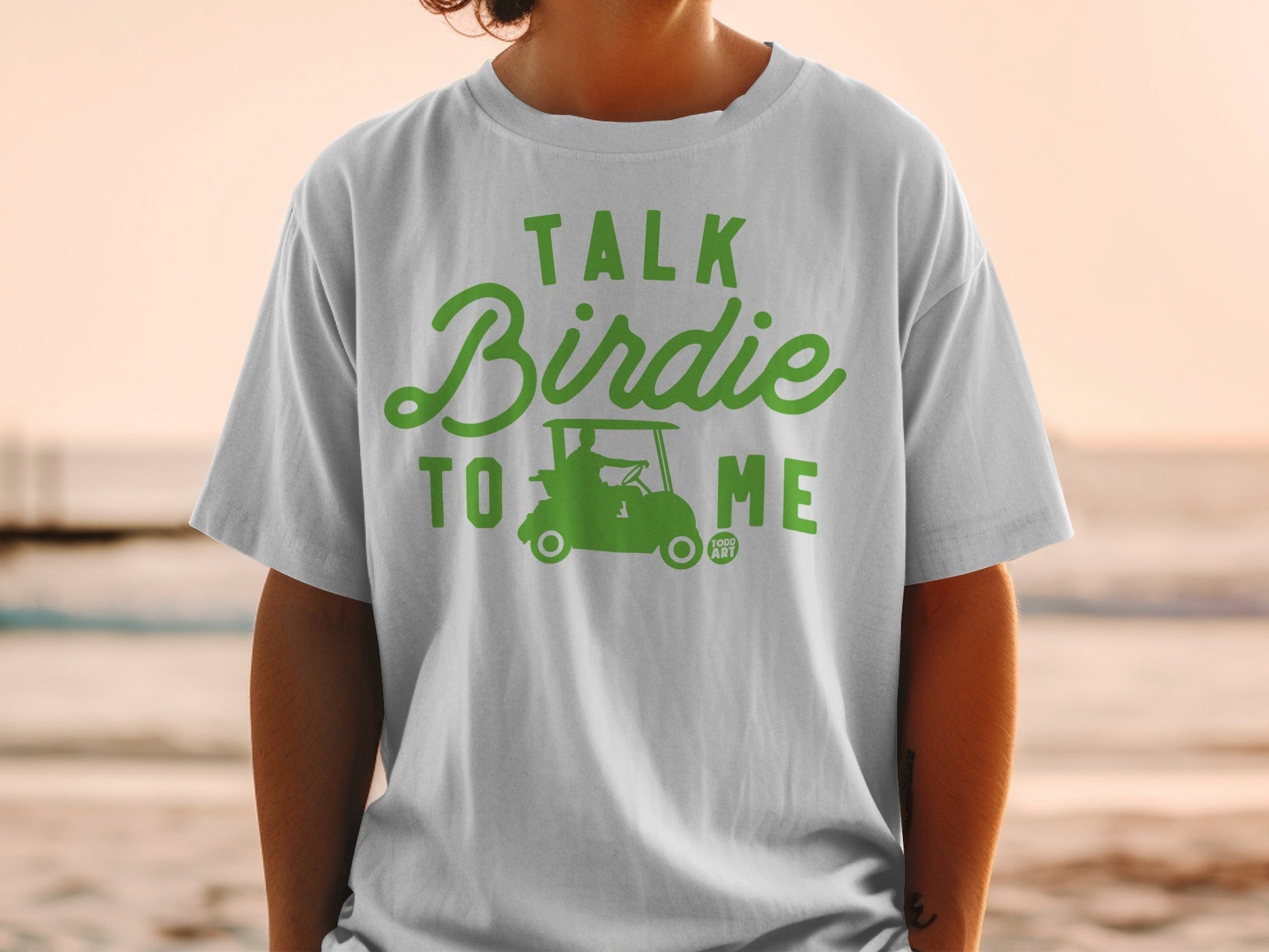 Green text "Talk Birdie To Me" with golf cart graphic on a white t-shirt.