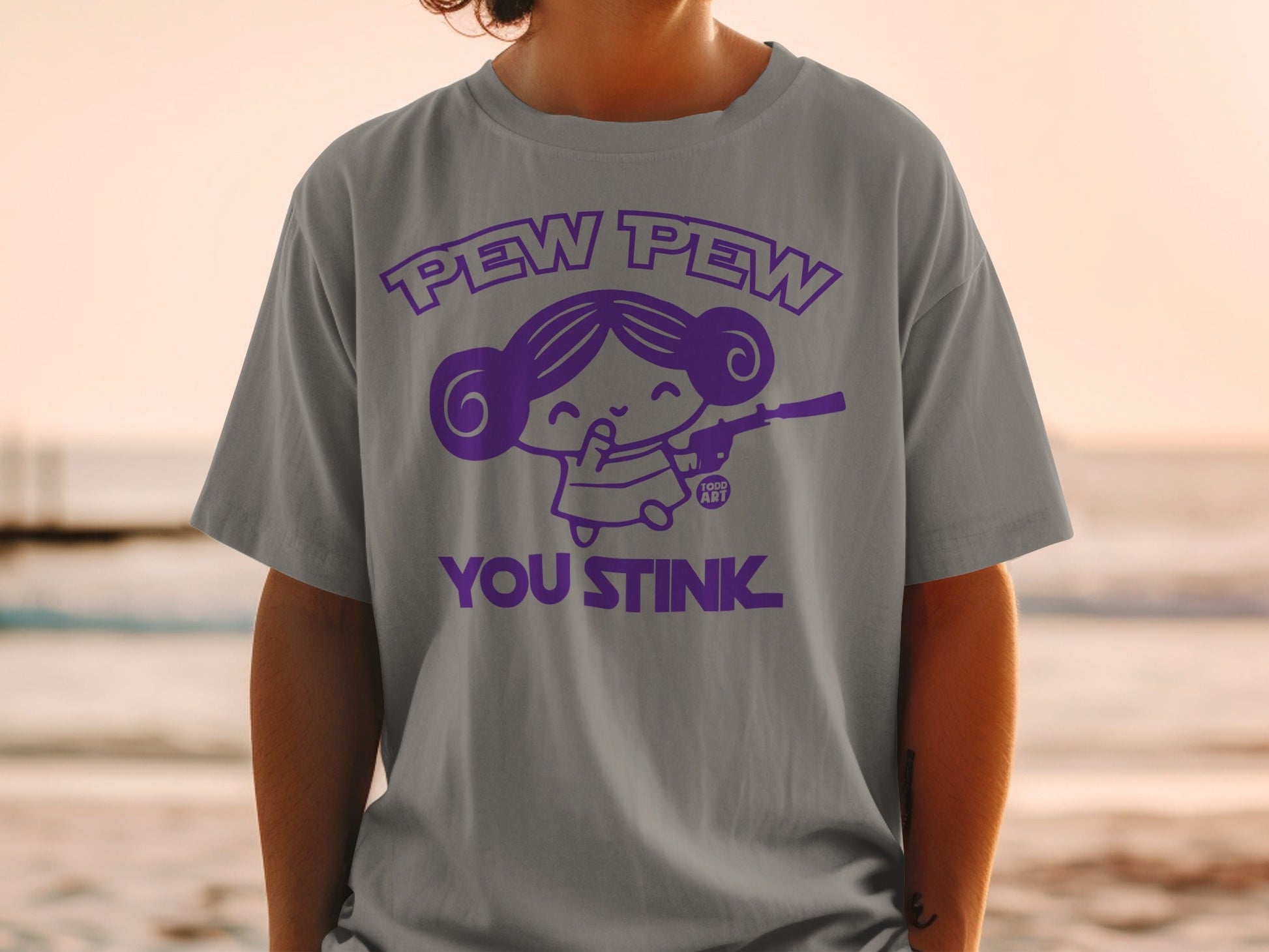 Grey t-shirt with purple graphic of cartoon girl holding a gun that says "Pew Pew You Stink".
