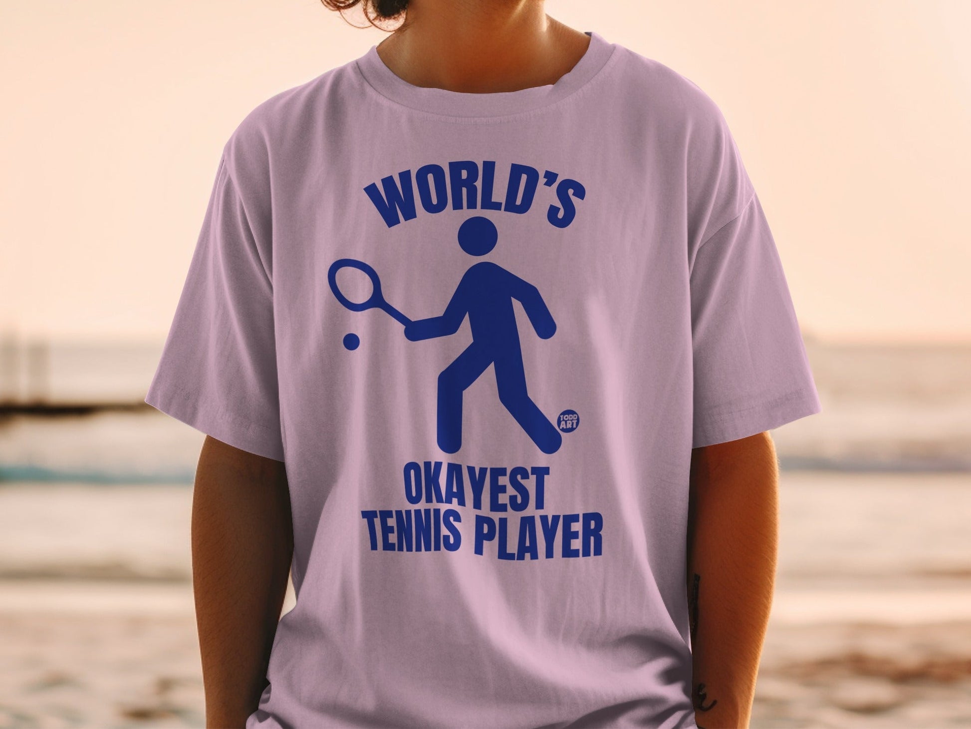 Purple t-shirt with blue graphic of a tennis player and text "WORLD'S OKAYEST TENNIS PLAYER".