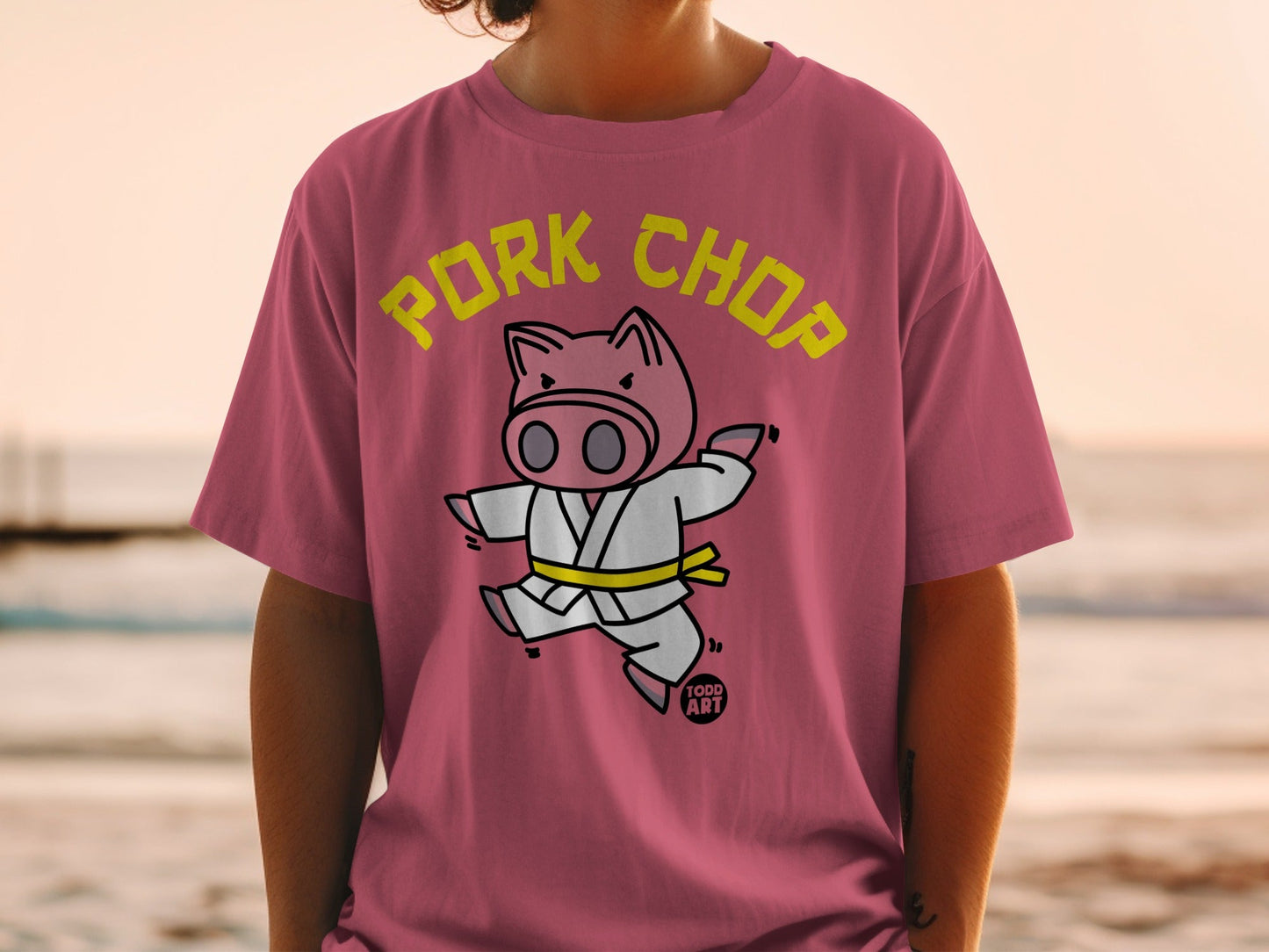 Cartoon pig wearing a yellow belt in a karate stance on a pink t-shirt.