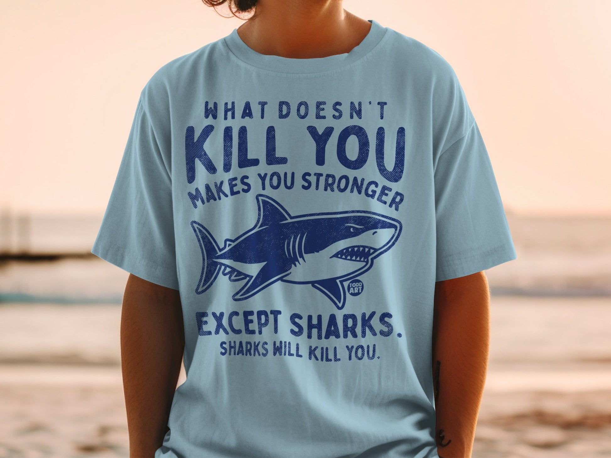 Light blue t-shirt with shark and text "What doesn't kill you makes you stronger, except sharks. Sharks will kill you."