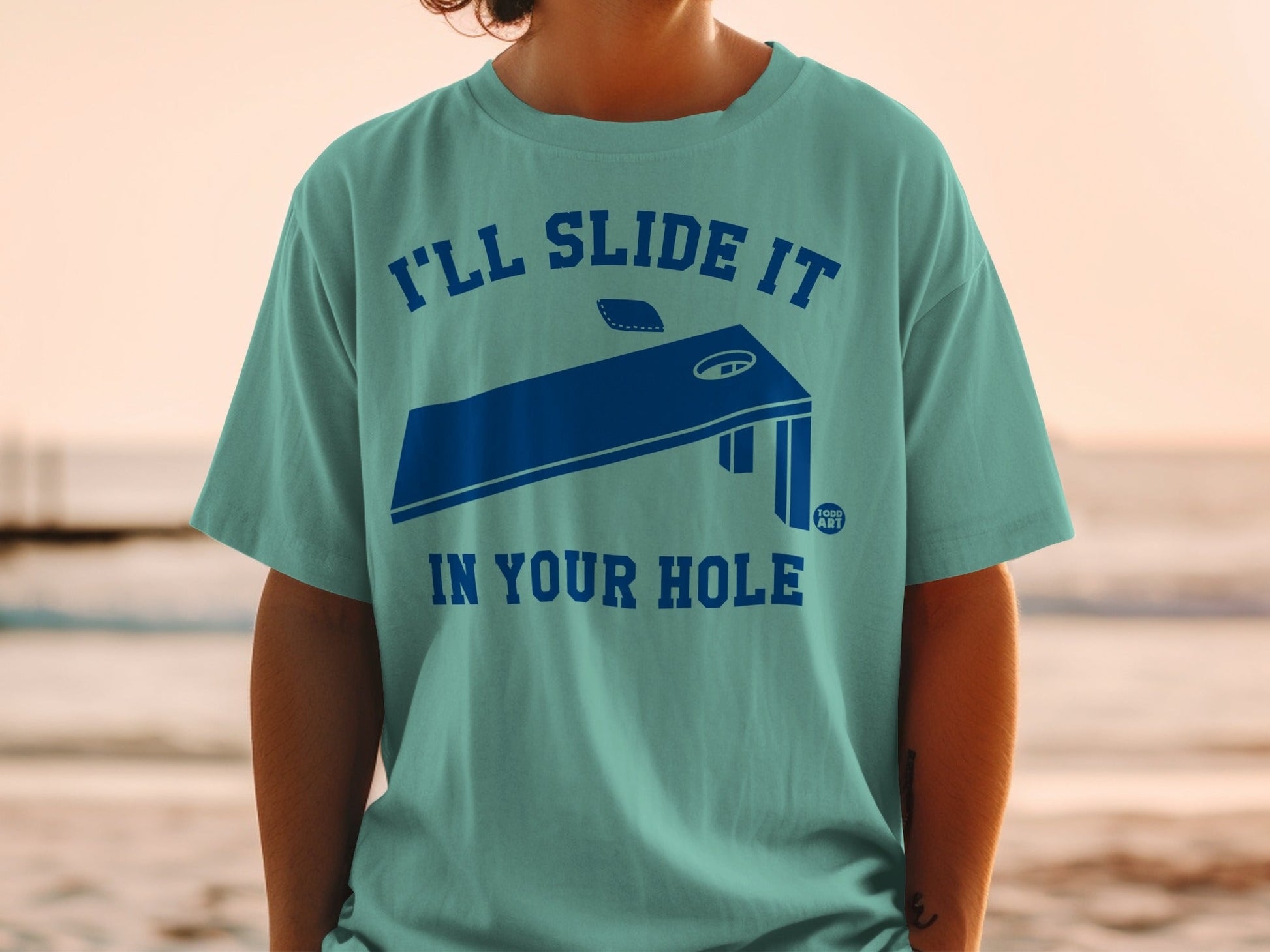Teal t-shirt with blue graphic of a cornhole board and text.