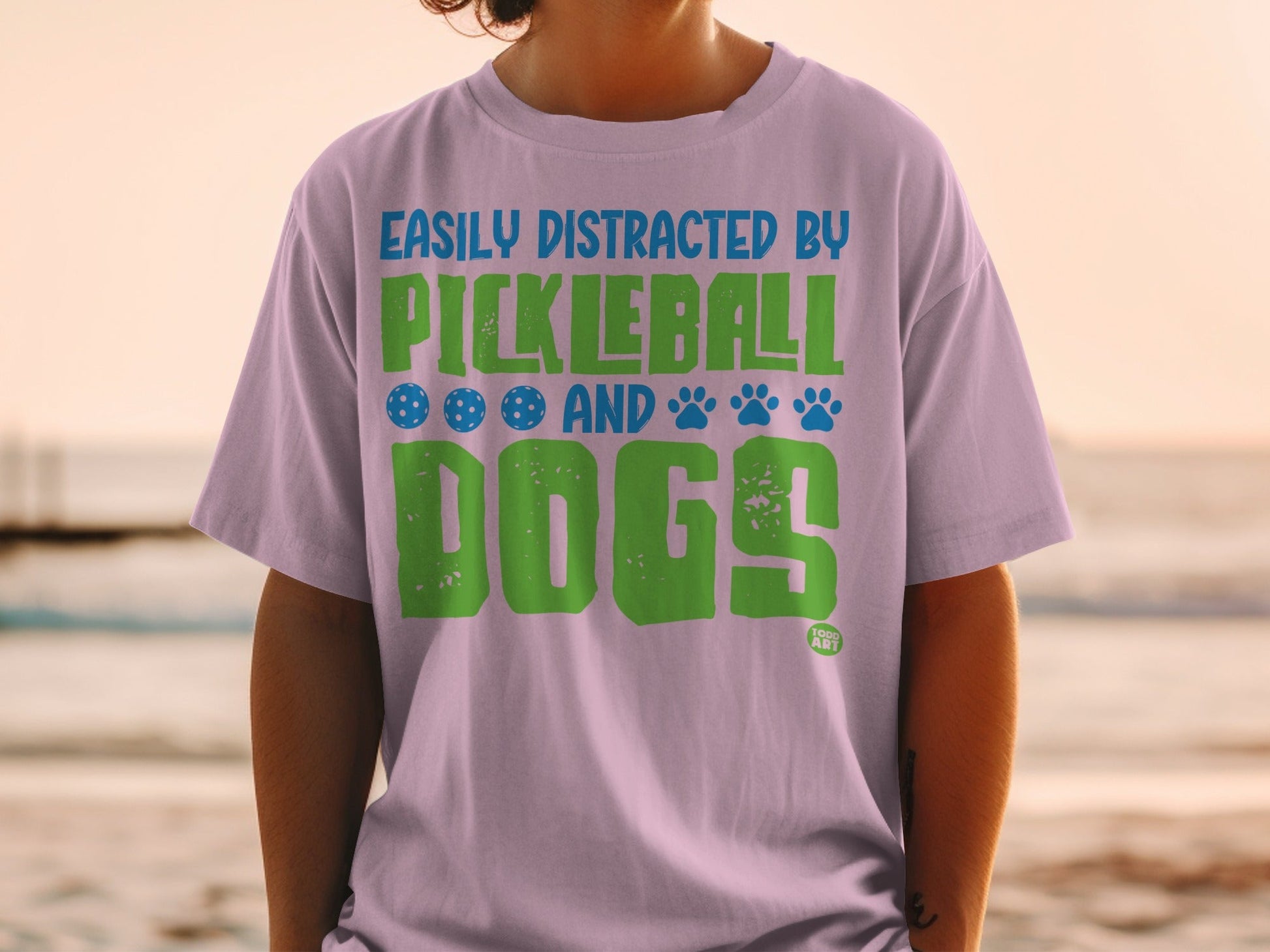 Light purple t-shirt with text "Easily Distracted by Pickleball and Dogs" in green and blue.