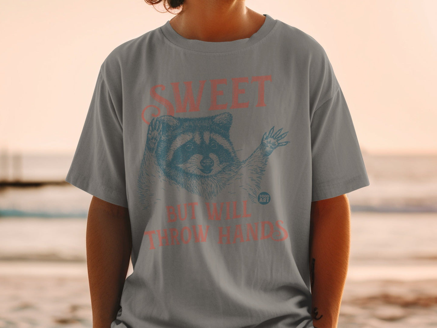 Graphic t-shirt featuring a raccoon and text: "SWEET BUT WILL THROW HANDS".