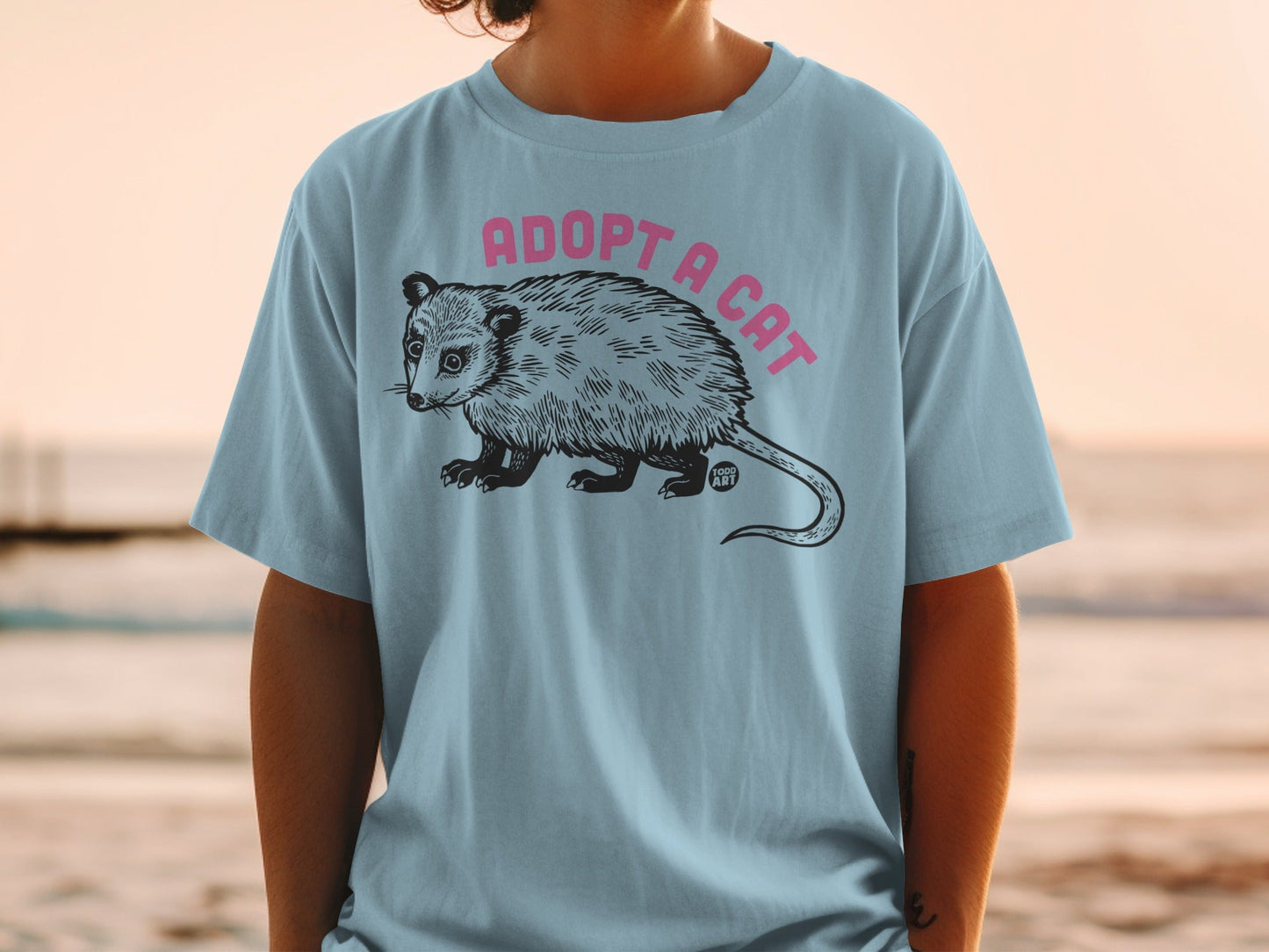 Light blue t-shirt featuring a black and white illustration of an opossum with "ADOPT A CAT" in pink text above.