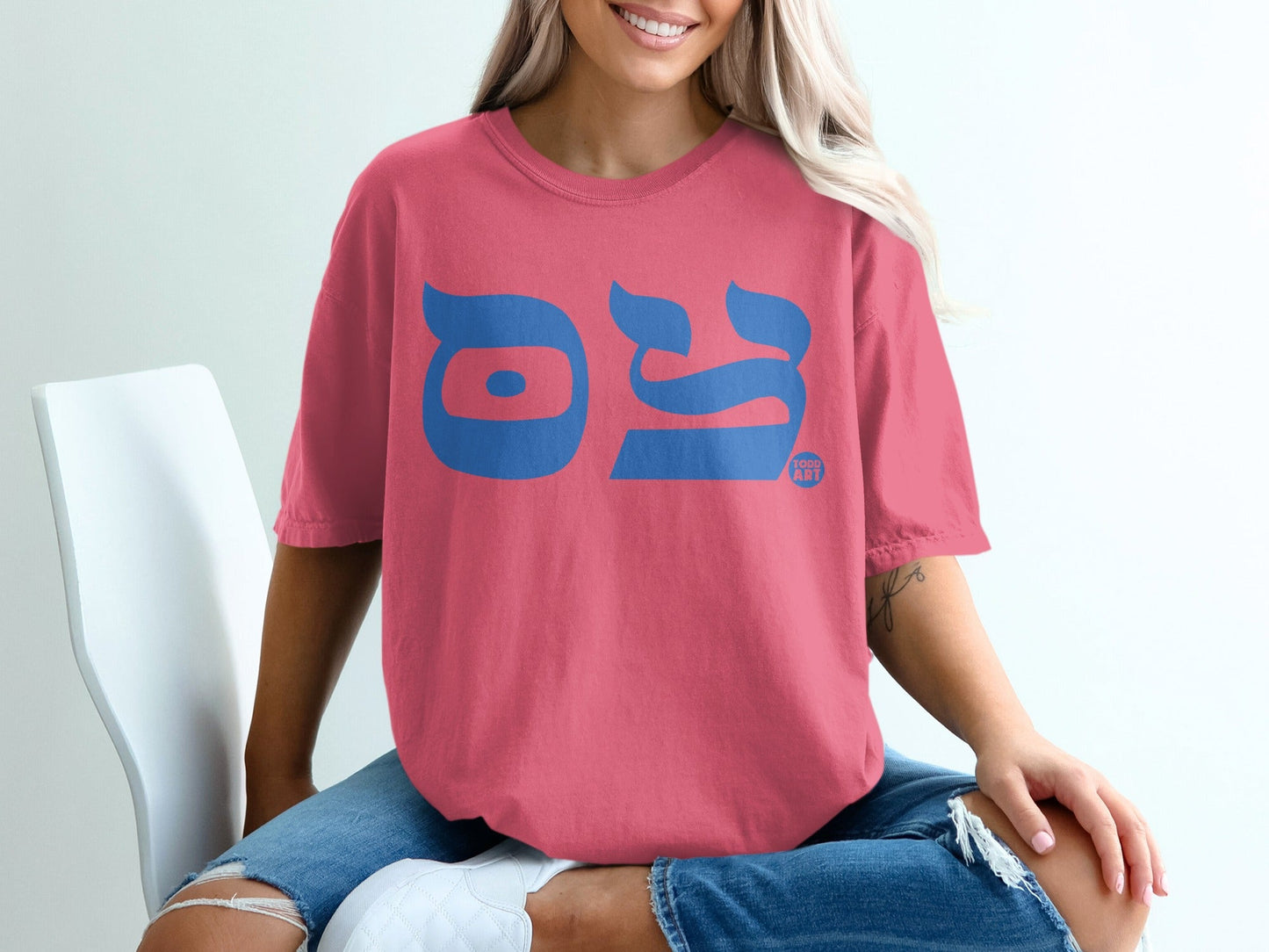 Woman wearing pink t-shirt with blue Hebrew text, seated on white chair.