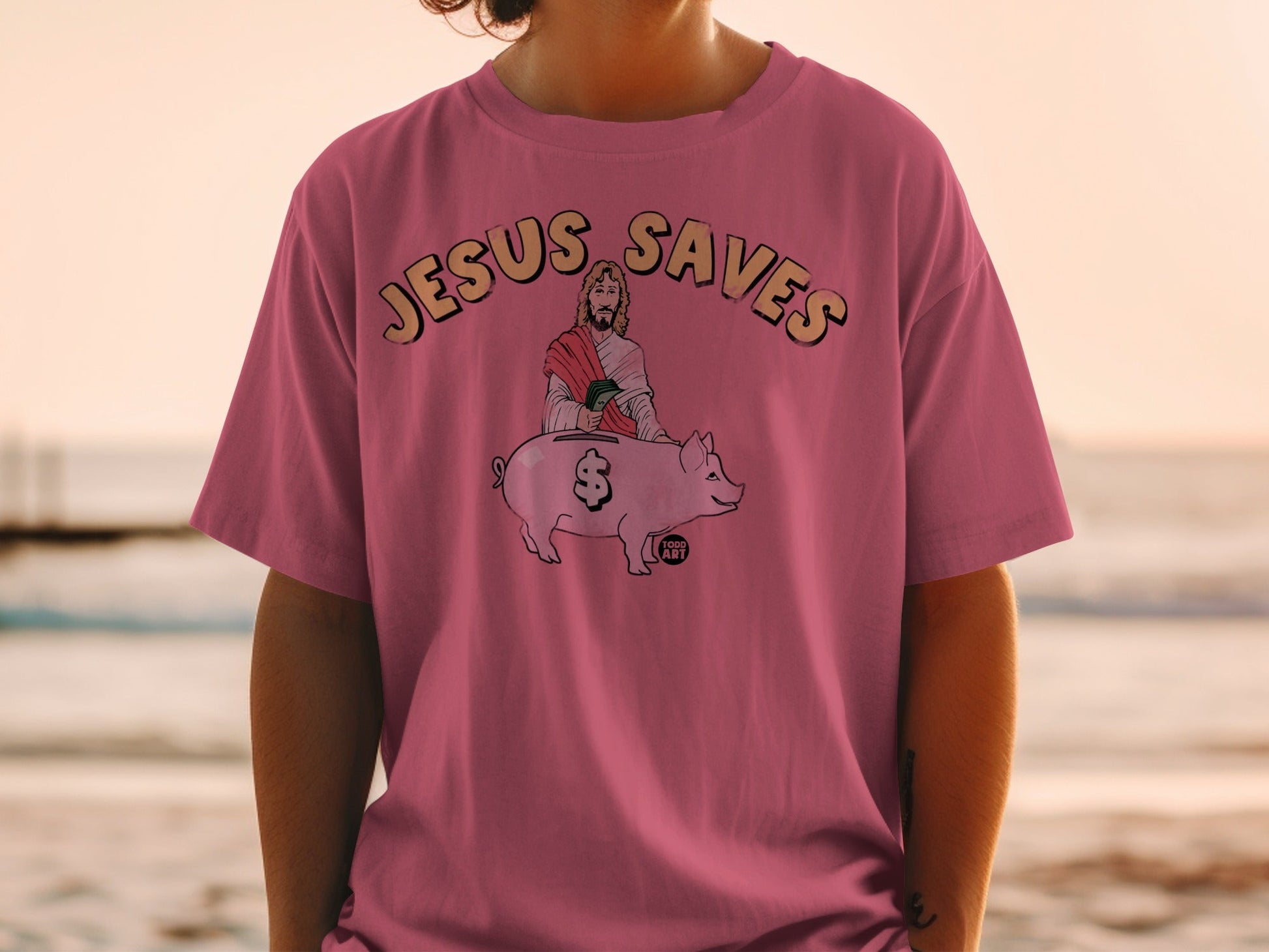 Pink t-shirt with "Jesus Saves" text and illustration of Jesus hugging a piggy bank.