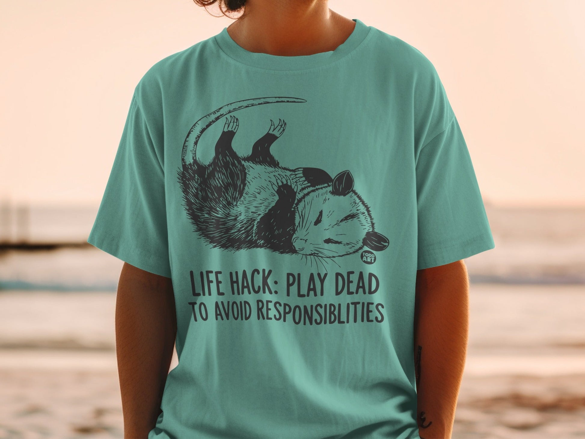 Teal t-shirt with opossum graphic and text "LIFE HACK: PLAY DEAD TO AVOID RESPONSIBILITIES".