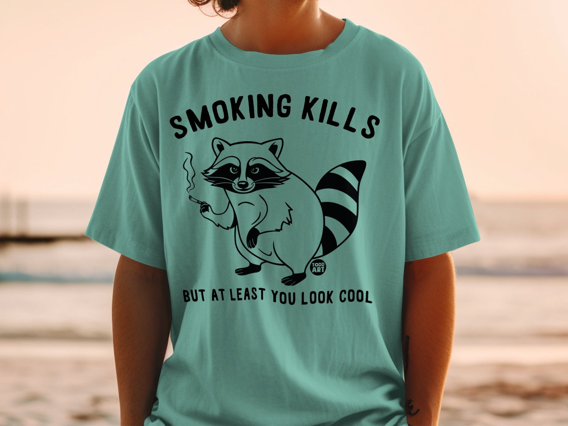 Teal t-shirt with a smoking raccoon graphic and the text "SMOKING KILLS BUT AT LEAST YOU LOOK COOL".