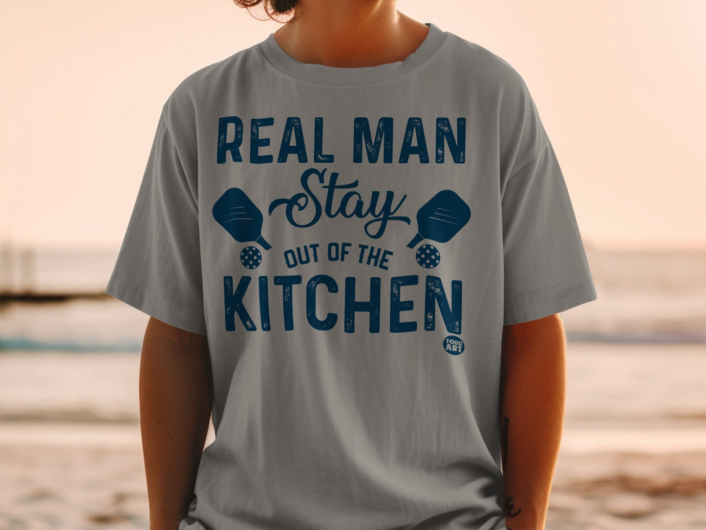 Grey t-shirt with blue text reading "REAL MAN STAY OUT OF THE KITCHEN" and pickleball paddles.